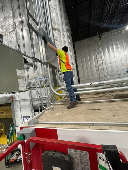 Worker wearing a yellow safety vest and gloves installing or inspecting electrical or HVAC wiring on a metal wall in an industrial or warehouse setting.