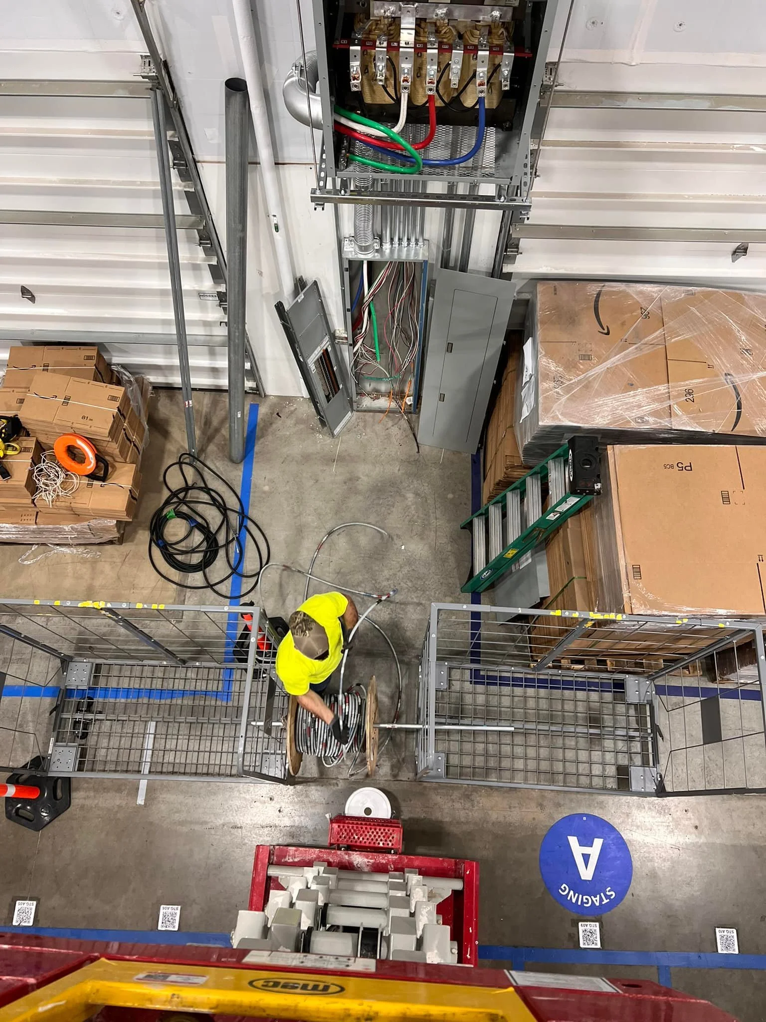 An overhead view of a worker in a yellow shirt wiring electrical panels in a warehouse, surrounded by pallets and tools.