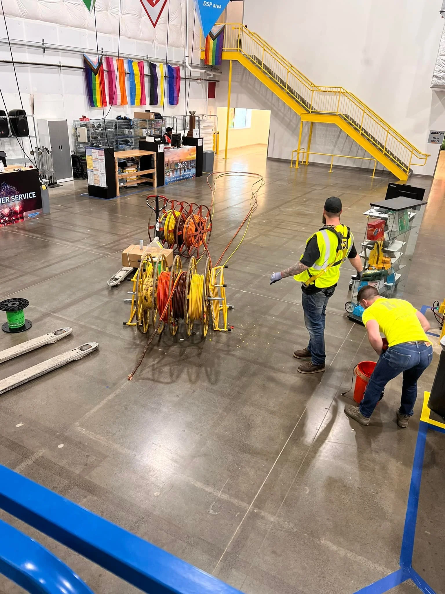 Workers setting up electrical cables on the floor of an indoor event space, with colorful flags hanging on the wall and a staircase in the background.