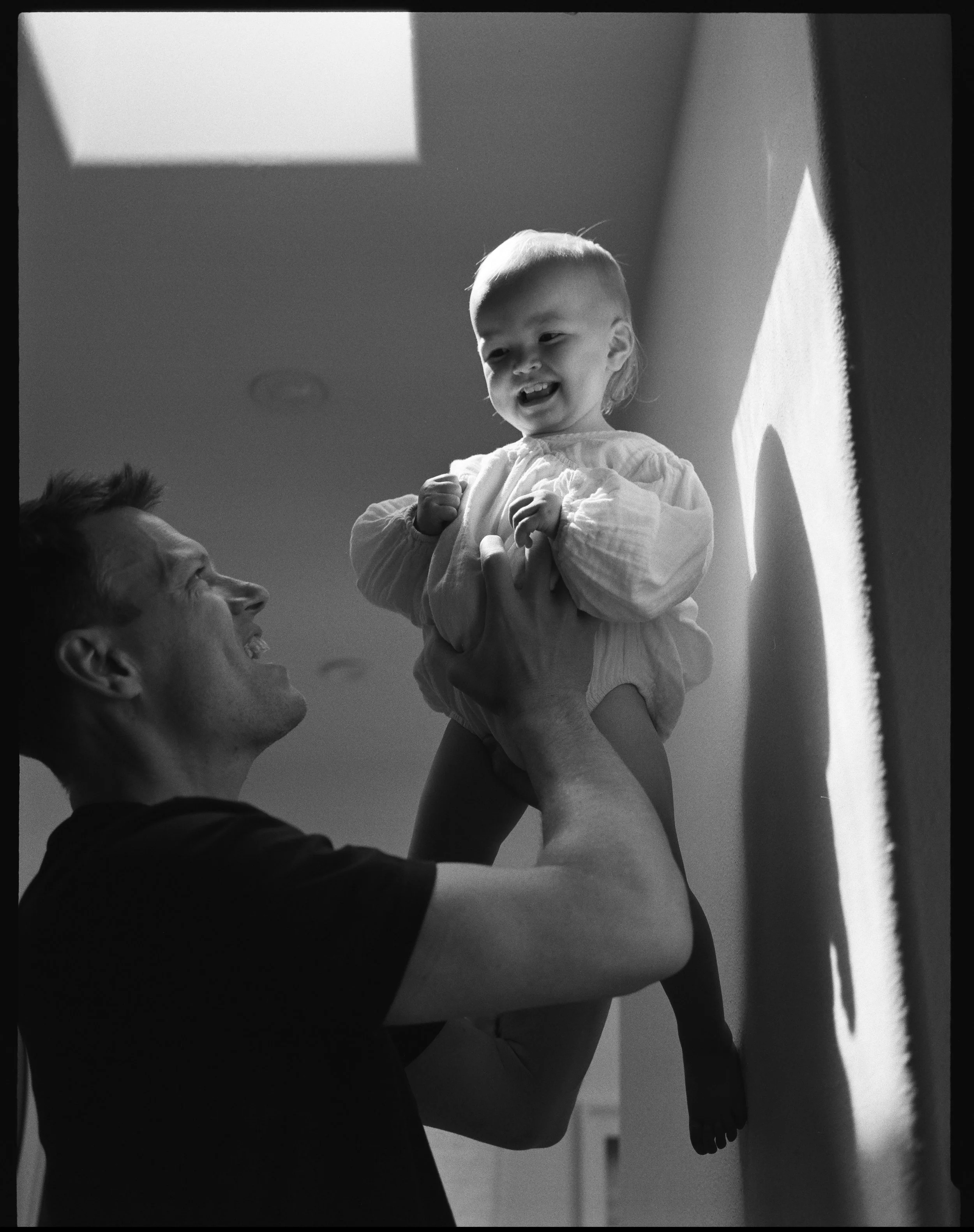 a medium format black and white film photo of a father holding up his infant daughter into a patch of light thats shining on the wall in front of them in their home in Sausalito 