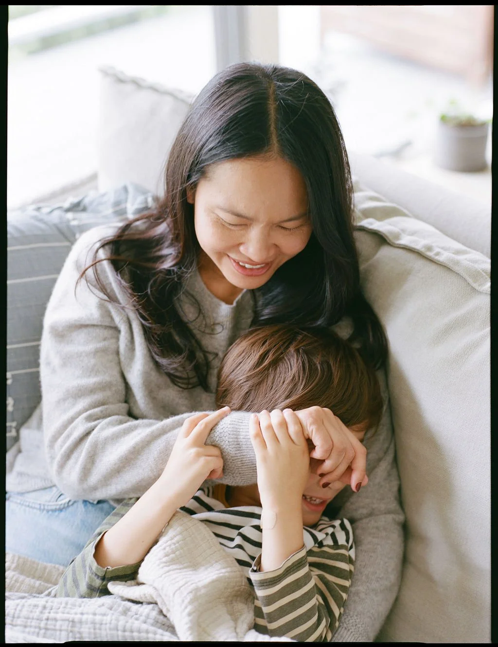A woman and a boy are sitting on a sofa, playing a game of hide and seek, laughing and having fun. Color medium format film image taken in Sausalito, California
