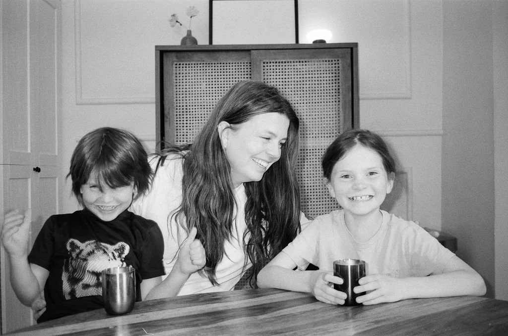 A woman with long hair and two young girls laughing and sitting at a table in a kitchen, holding mugs.