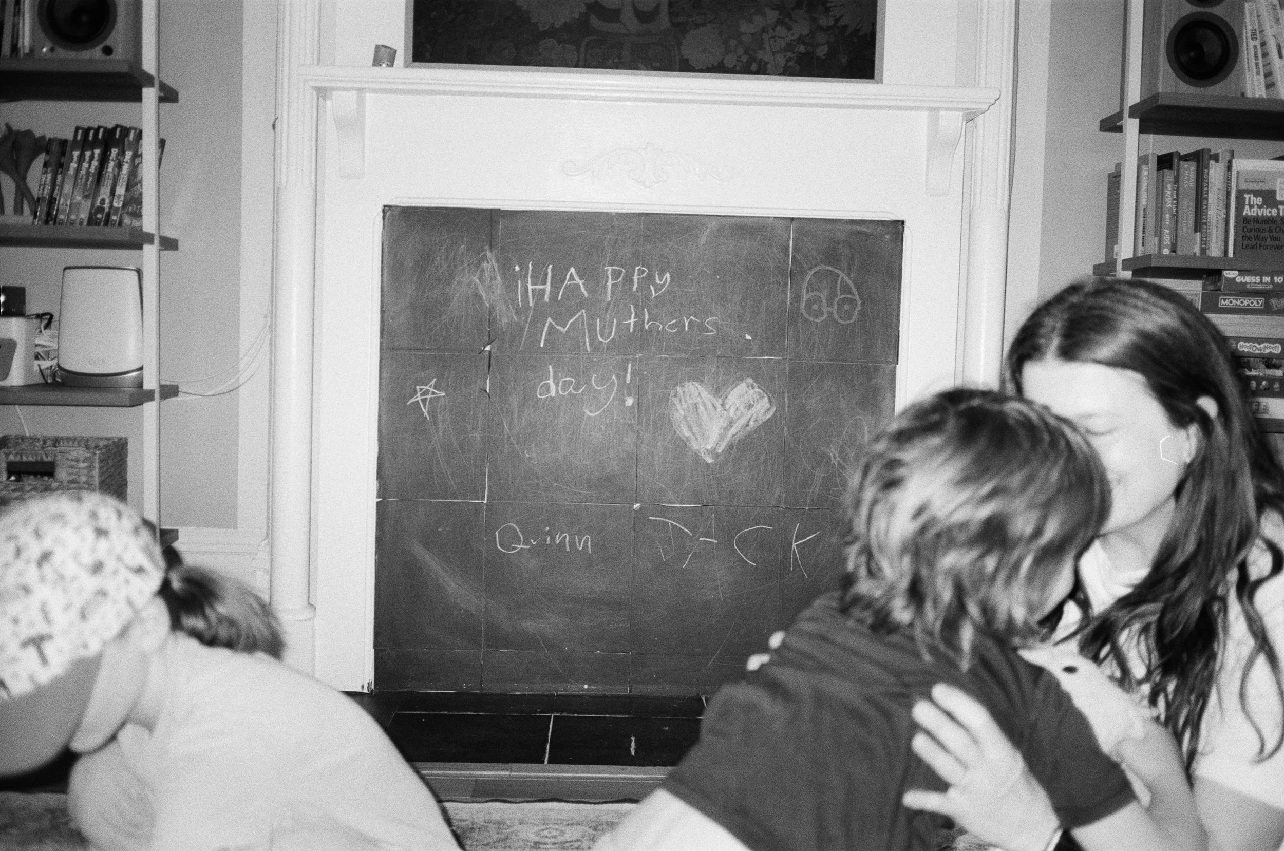 A black and white photo of a living room with children, a fireplace with a chalkboard above it, and shelves with books and a speaker. The chalkboard has happy muther's day, with a heart and star drawn on it.