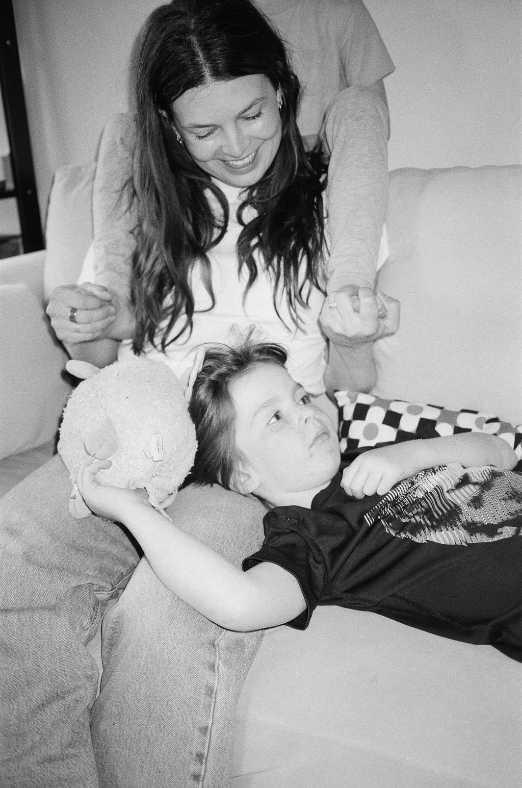 A woman playfully clenches her fists and smiles while a young girl lies on a couch holding a plush toy. The girl appears thoughtful and relaxed.