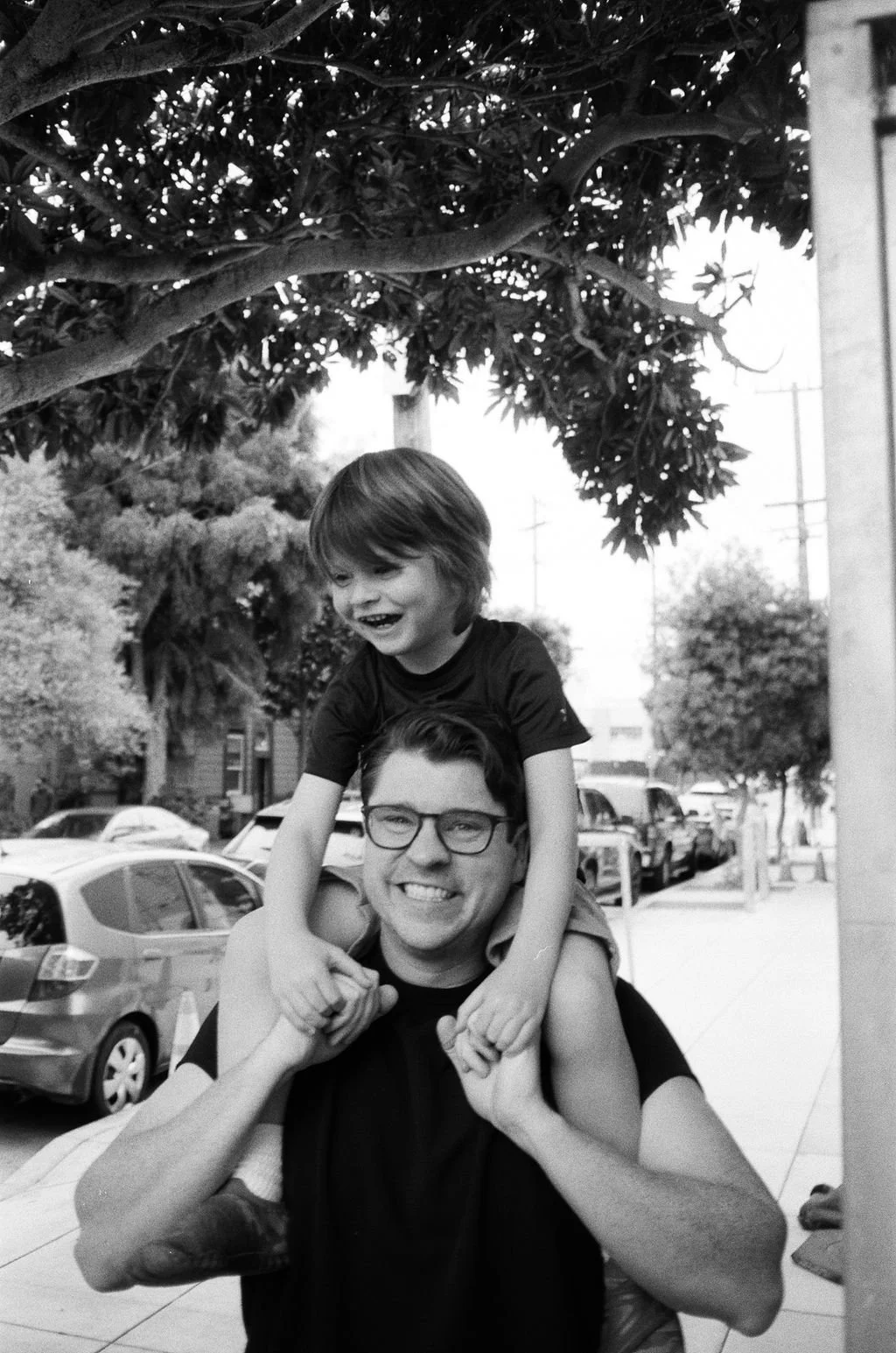 A man with glasses carrying a smiling young boy on his shoulders, outside on a street with parked cars and trees, under a tree with dense foliage.