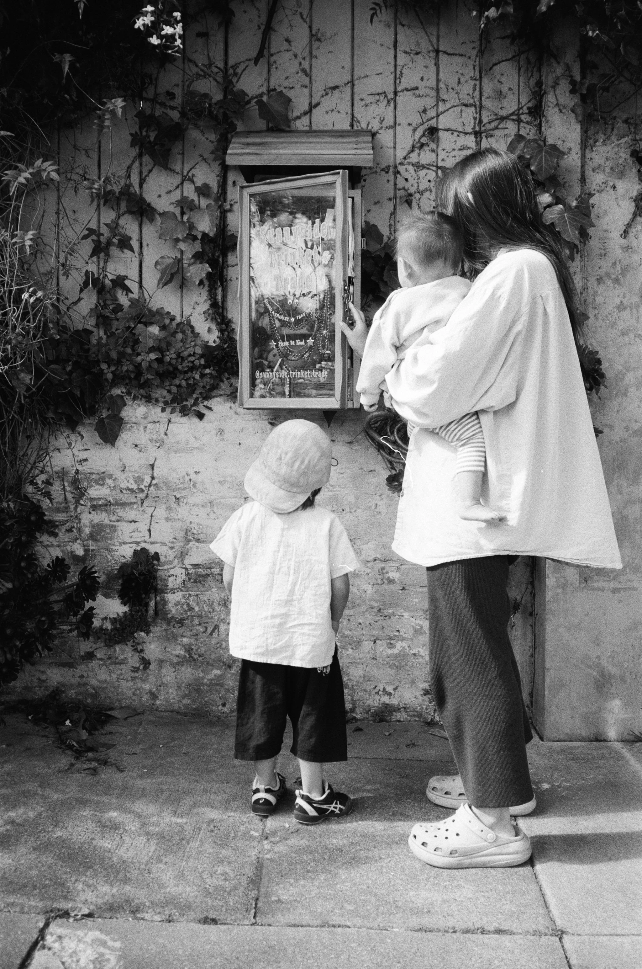 a black and white 35mm film photo of a mother holding her infant son and her toddler son standing next to her while they look at a trinket box on the side of a wall on a street in San Francisco, California 