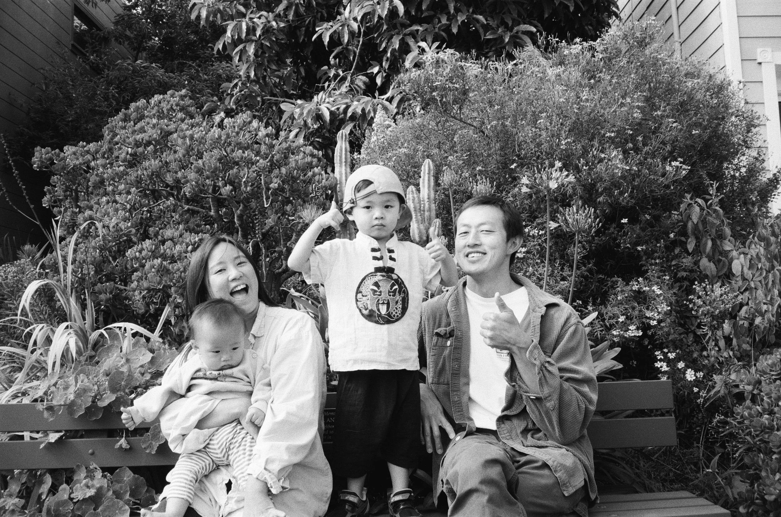 a 35mm black and white film image of an asian family consisting of a mother, father and two little boys sitting on a park bench in San Francisco, California. The father and the older son are holding their thumbs up, the mother is smiling and the infa
