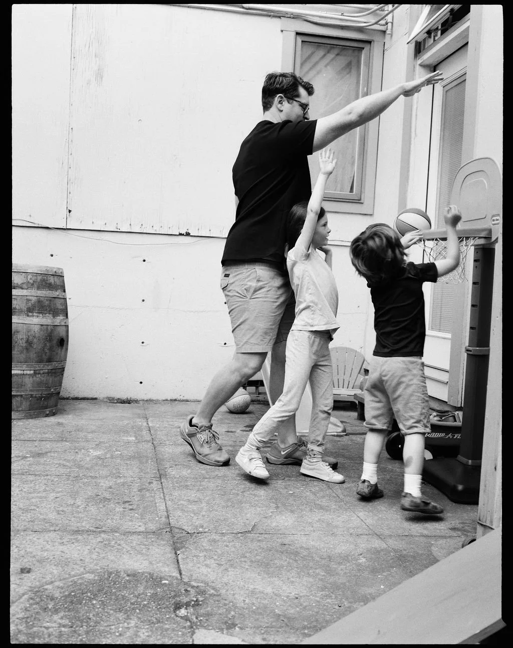 A man and two children playing basketball outdoors, with the man attempting a shot while the children defend or reach for the ball.