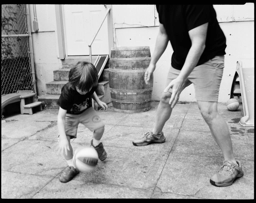 A young boy playing with a soccer ball while an adult crouches nearby in an outdoor backyard space.