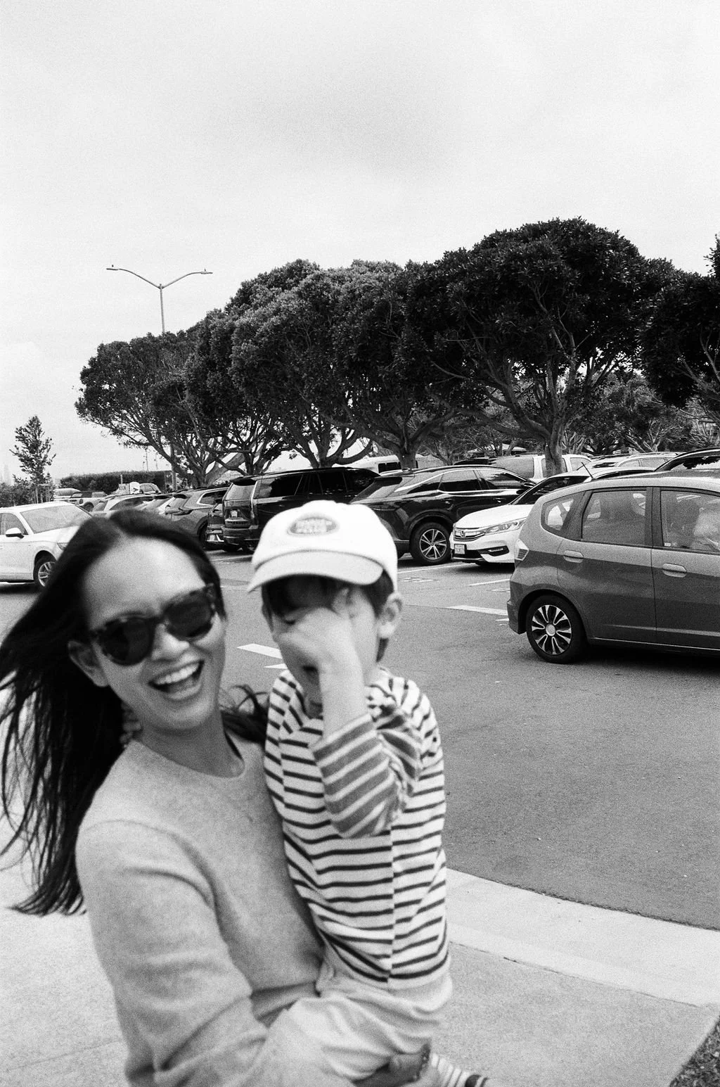 A woman wearing sunglasses and a sweater holding a young boy in a striped shirt and baseball cap, both smiling and enjoying outside in a parking lot with cars, trees, and a cloudy sky in the background. 35mm film photo taken in Sausalito, California.