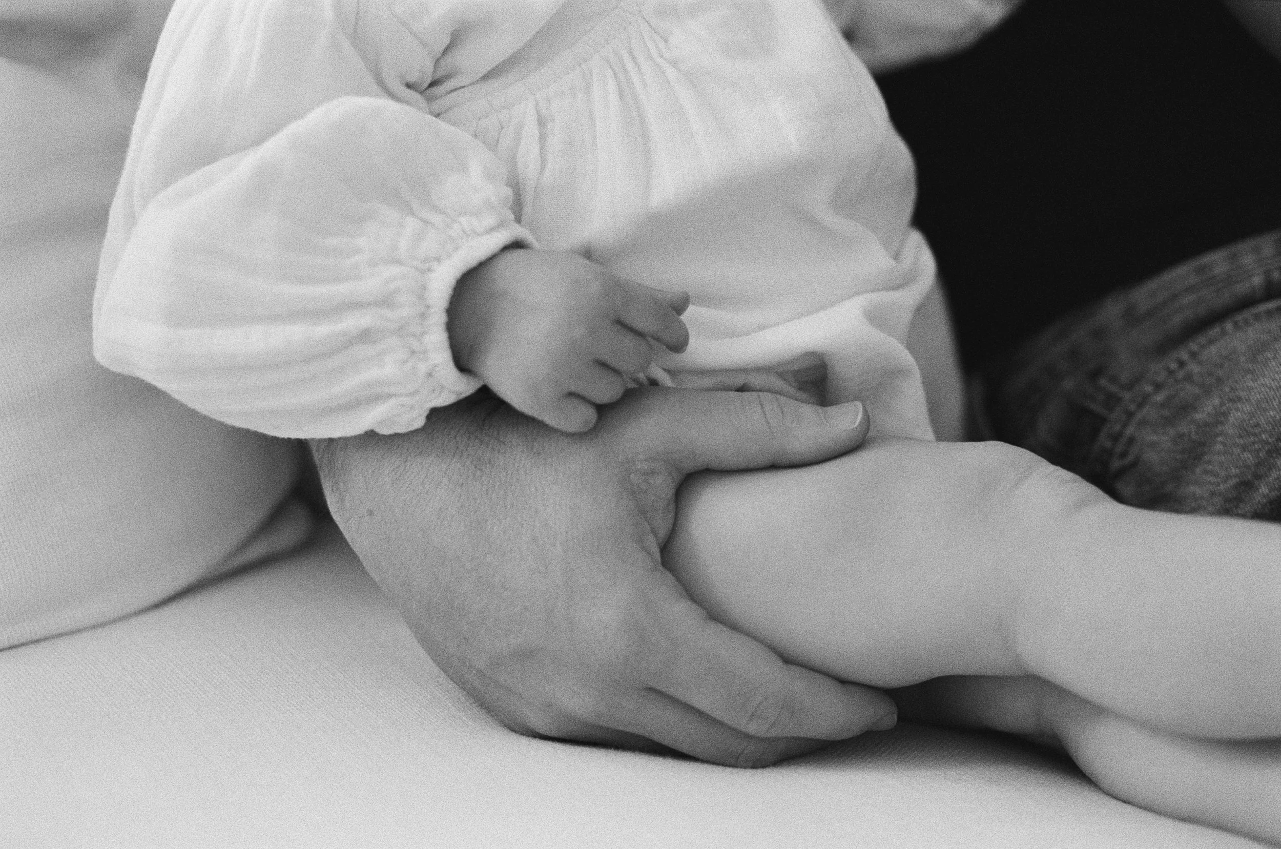black and white 35mm film image of a father holding the leg of his infant daughter while they sit on the couch in their home in Sausalito, California.
