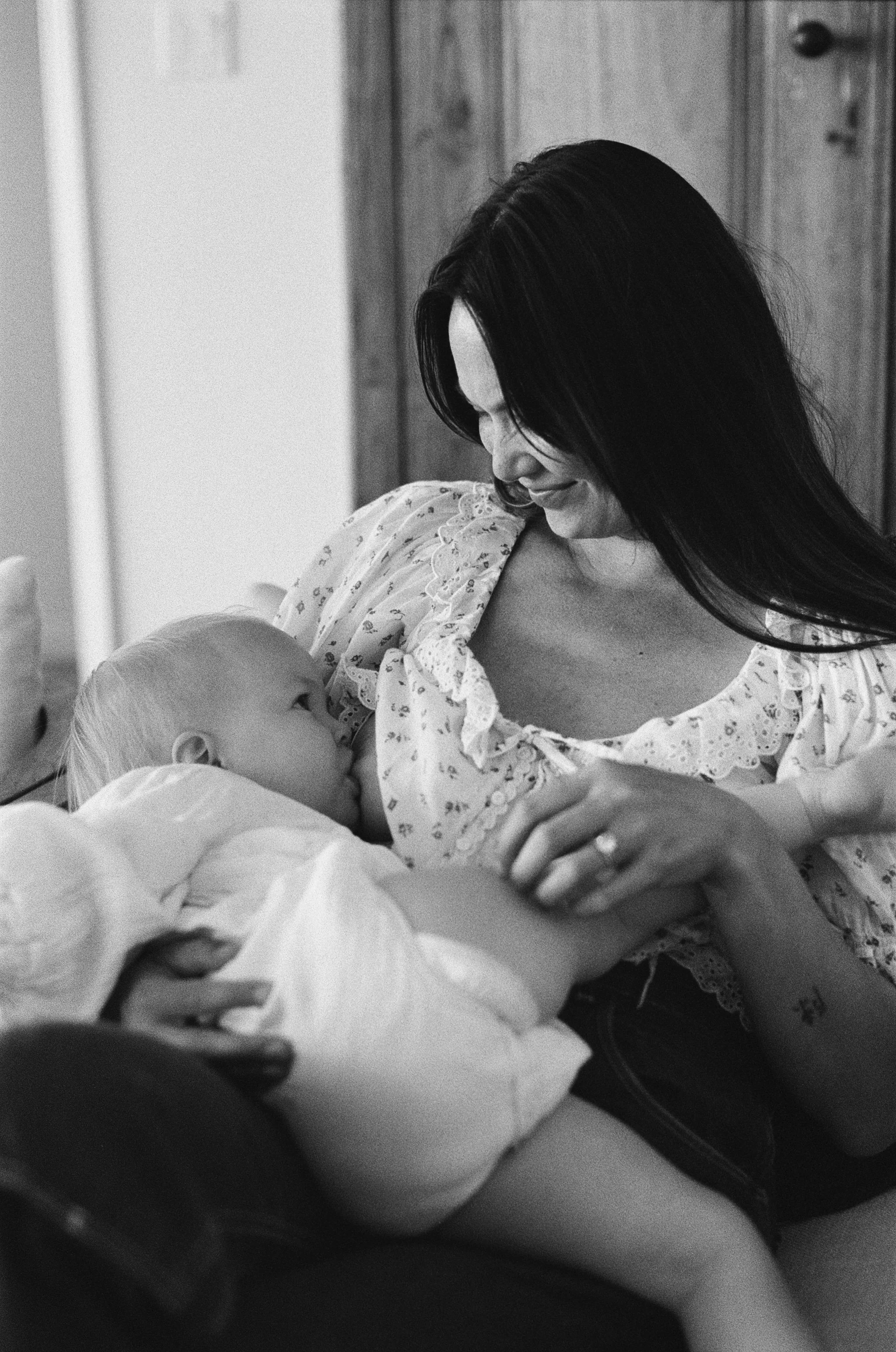 a black and white 35mm film photo of a mother sitting in a chair in her living room while she nursing her infant daughter at home in Sausalito, California 