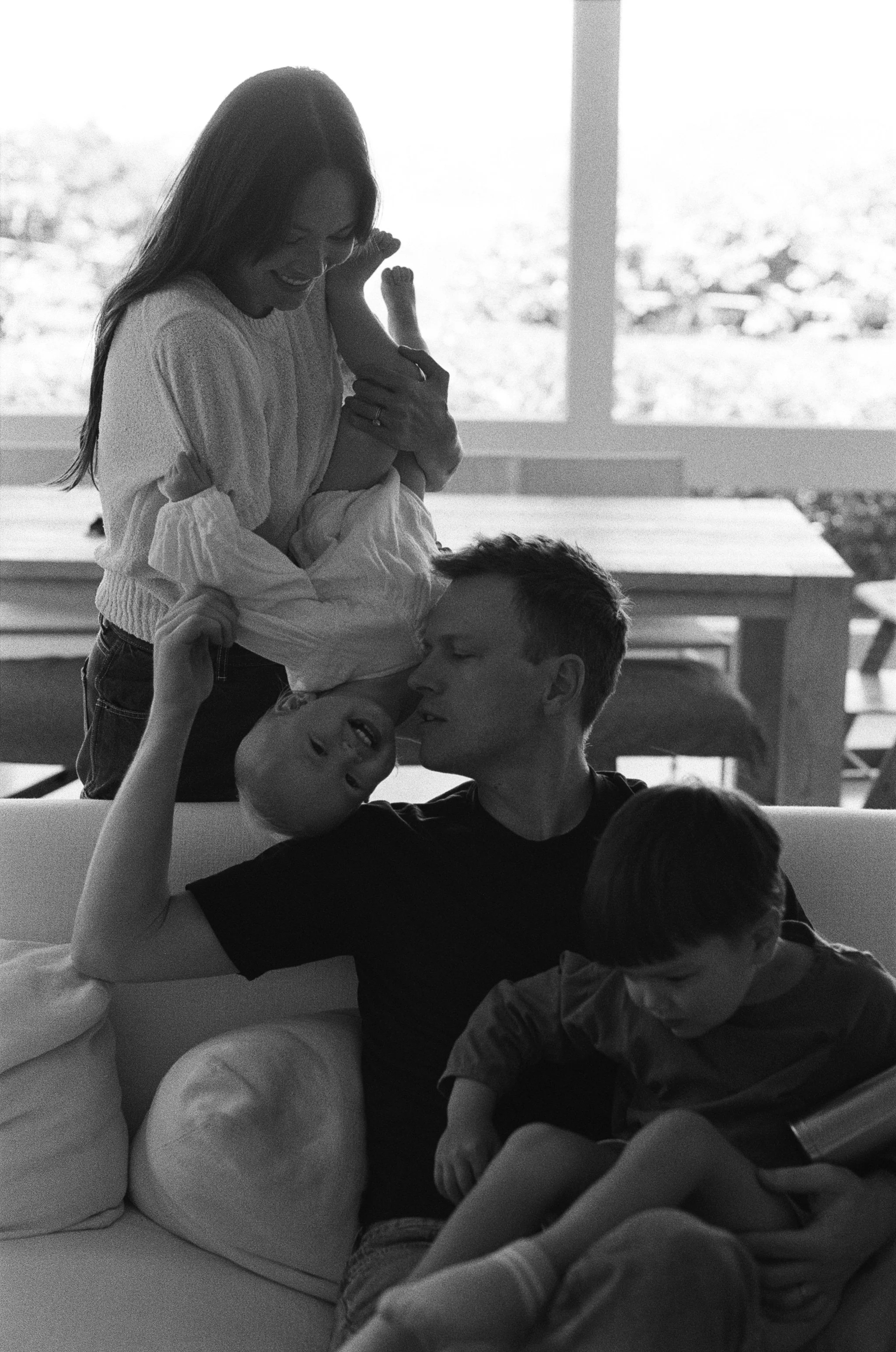 a black and white 35mm film image of a mother dangling her infant daughter playfully upside down while the father sits on the couch with their son in a tender family moment in their home in Sausalito, California 
