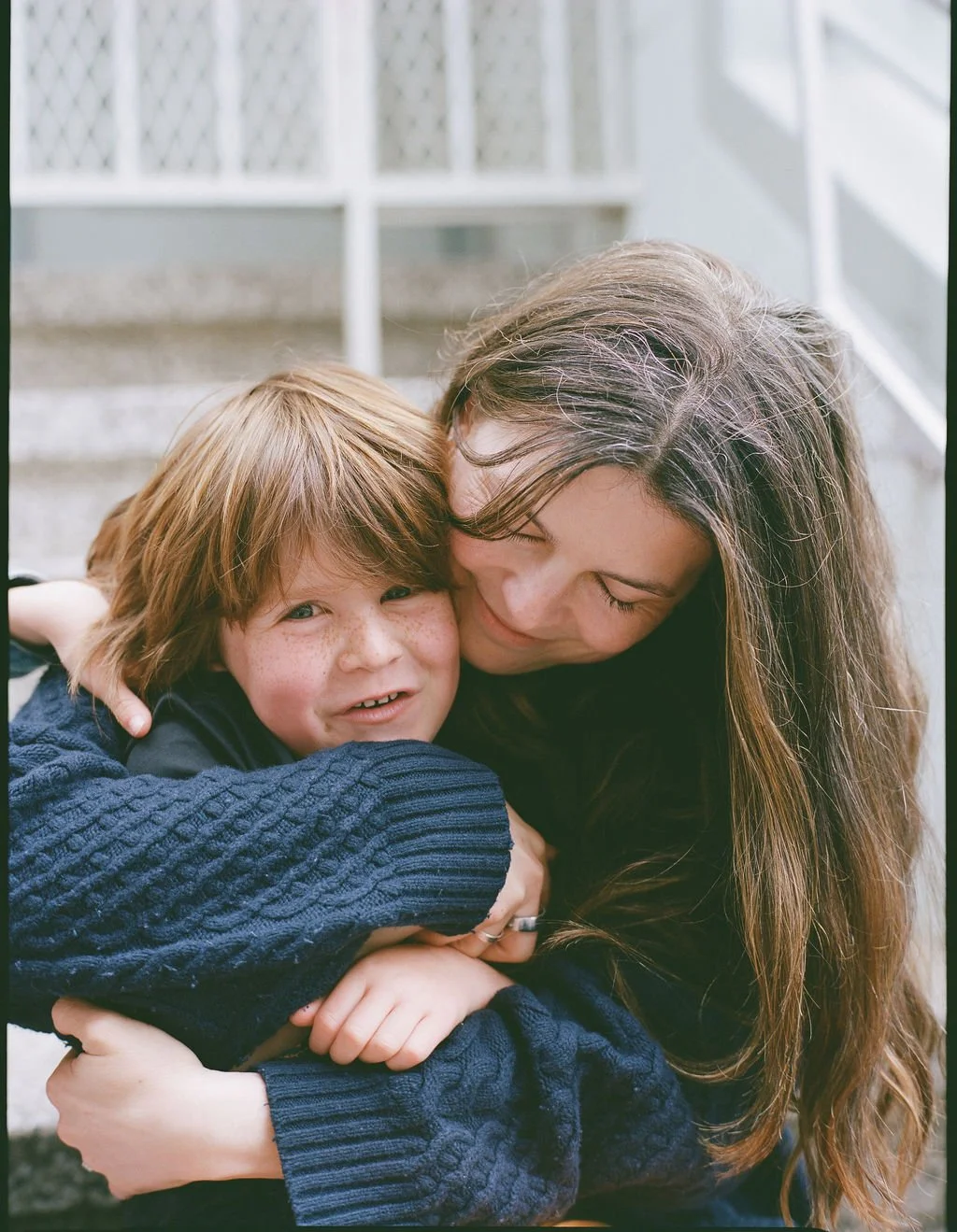 A woman hugging a young boy with red hair and freckles, both smiling indoors against a white lattice background.