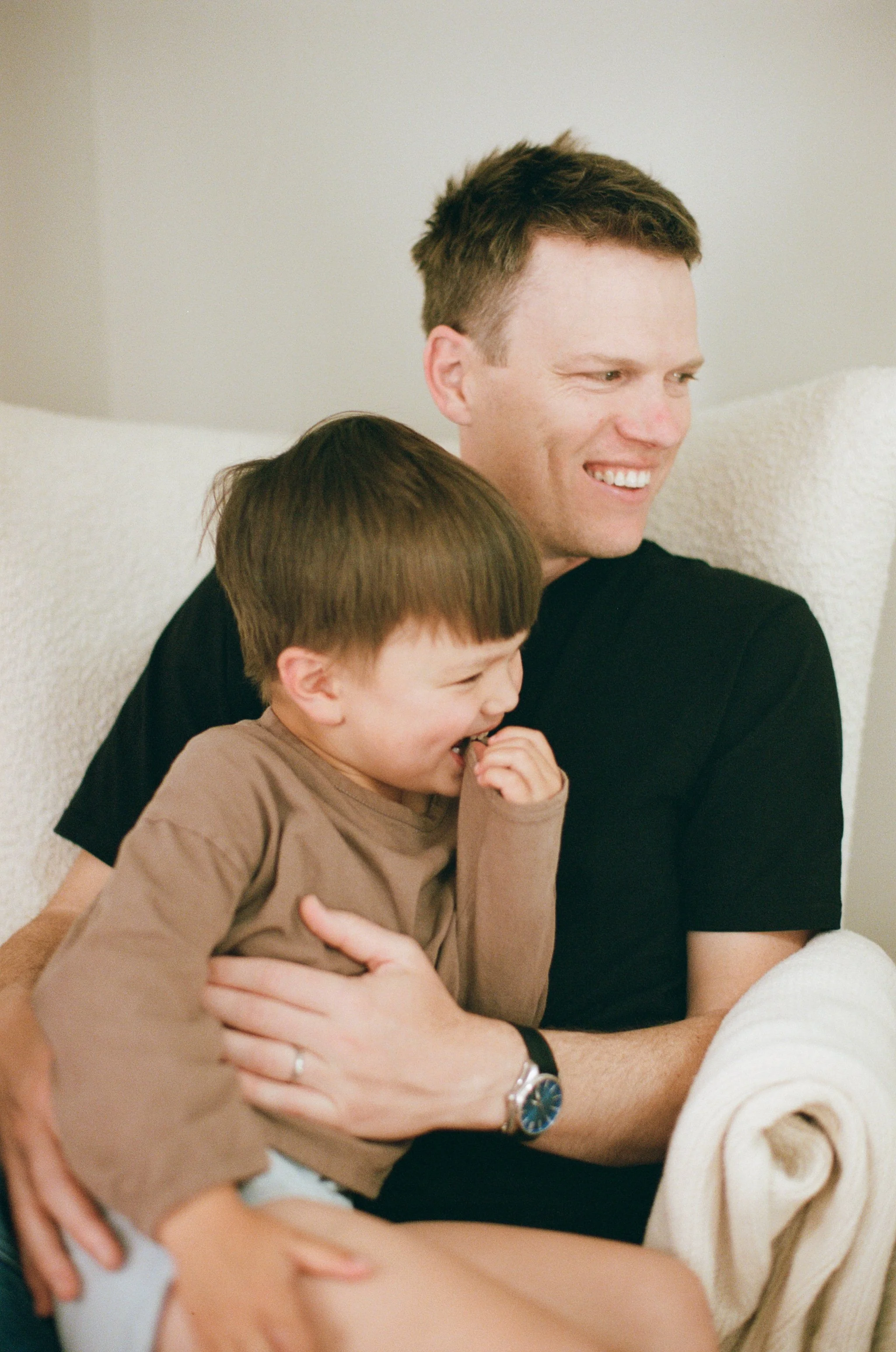 a color 35mm film photo of a father sitting on a chair with his son laughing in their home in Sausalito, California 
