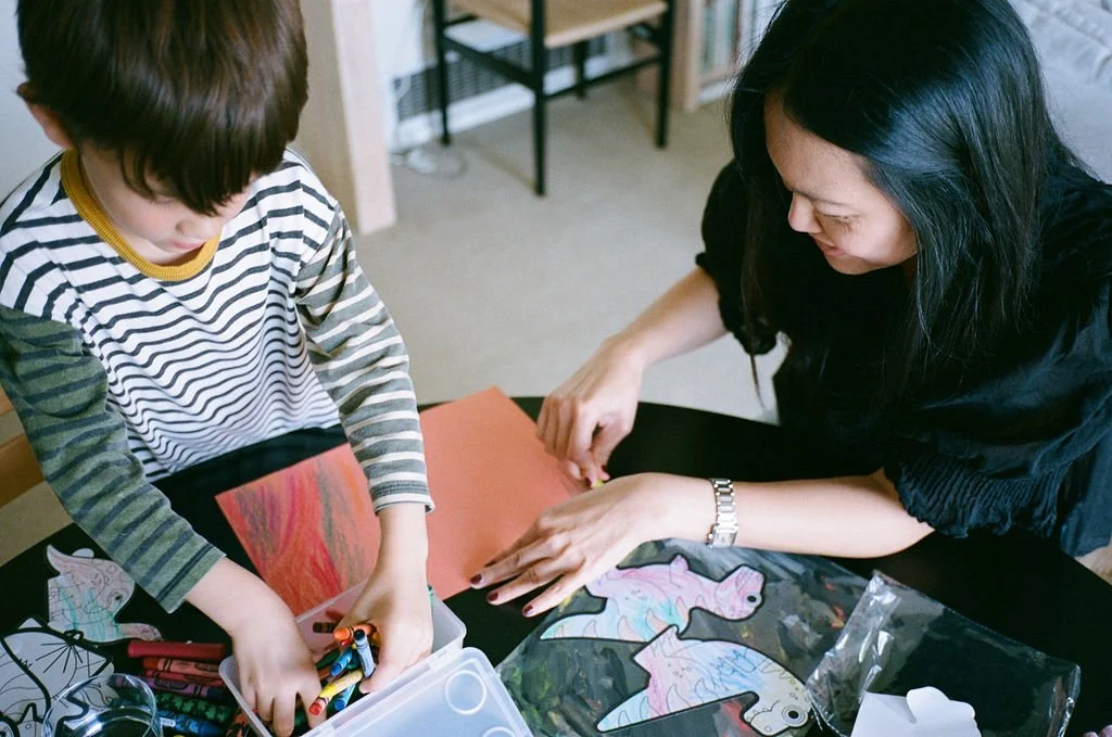 A woman and a young boy engaging in arts and crafts at a table. The boy is holding markers, and the woman is helping him with a project involving paper cutouts of animals.