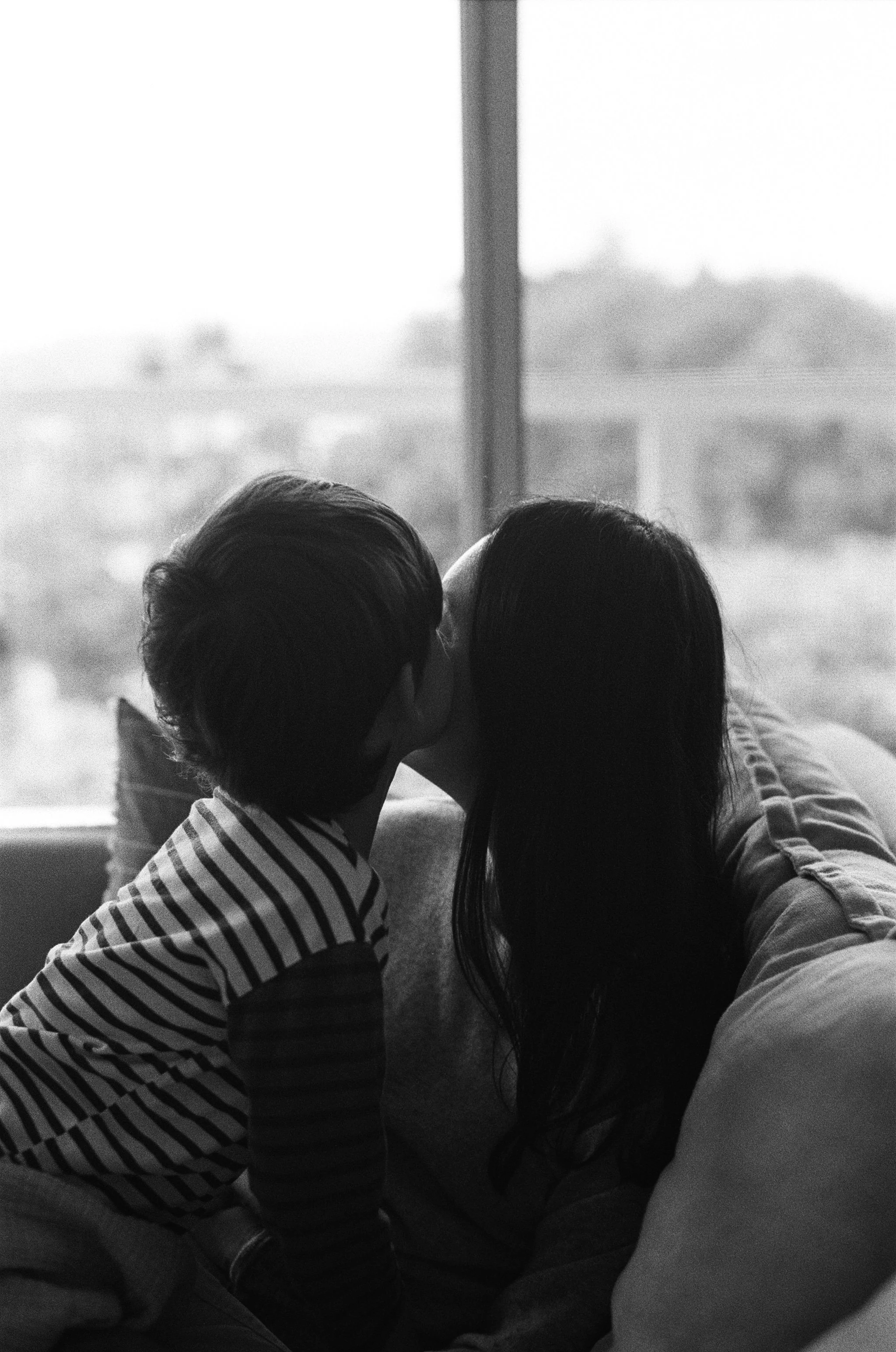 black and white 35mm film photo of a 5 year old son kissing the cheek of his mother. you cant see their faces, just the side of their faces. They are sitting on the couch in front of a large open window at their home in Sausalito, CA. 