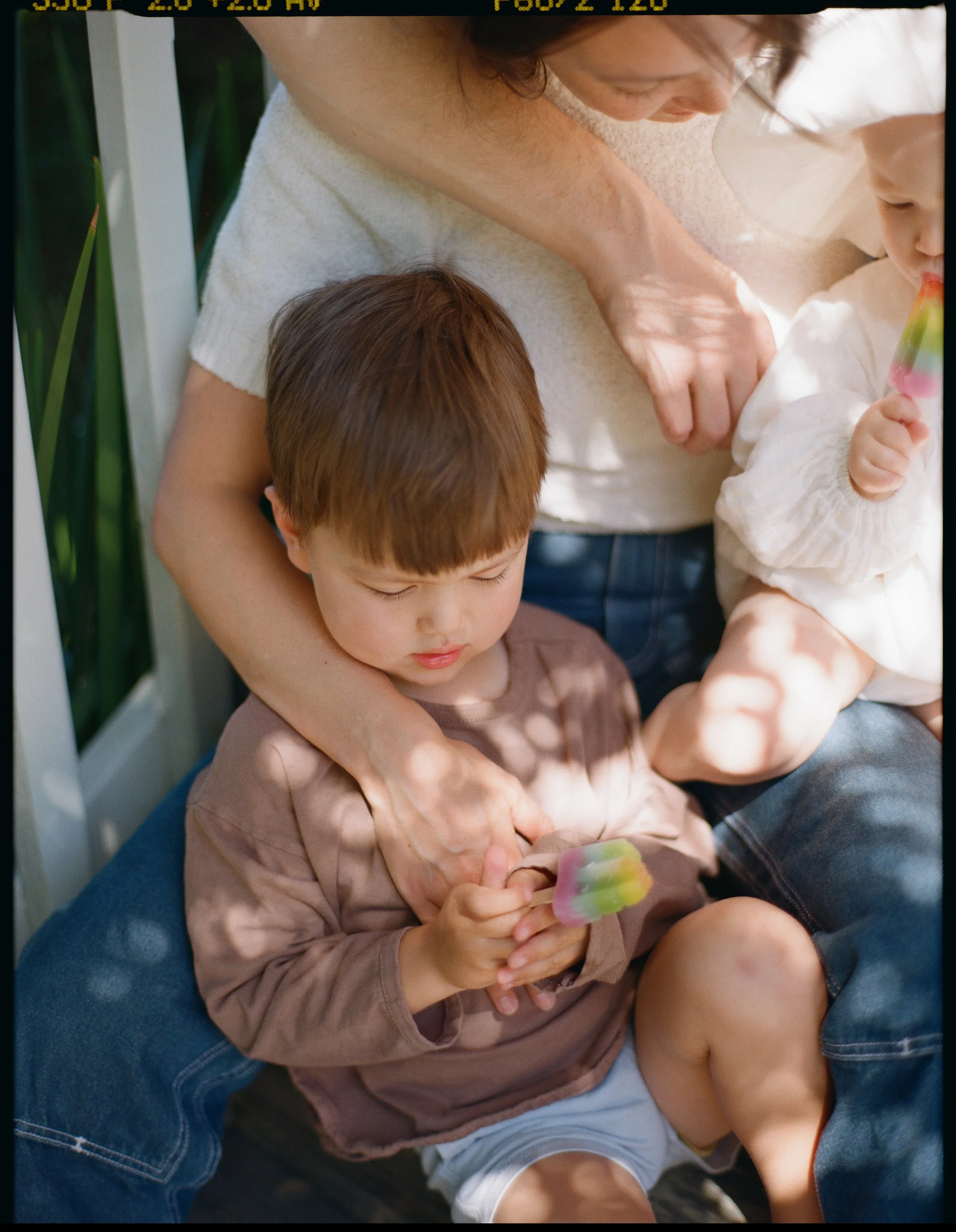a color medium format film image of a little boy sitting on the front steps of the porch of his home looking at a popsicle as he sits on his mother's nap in Sausalito, California. 