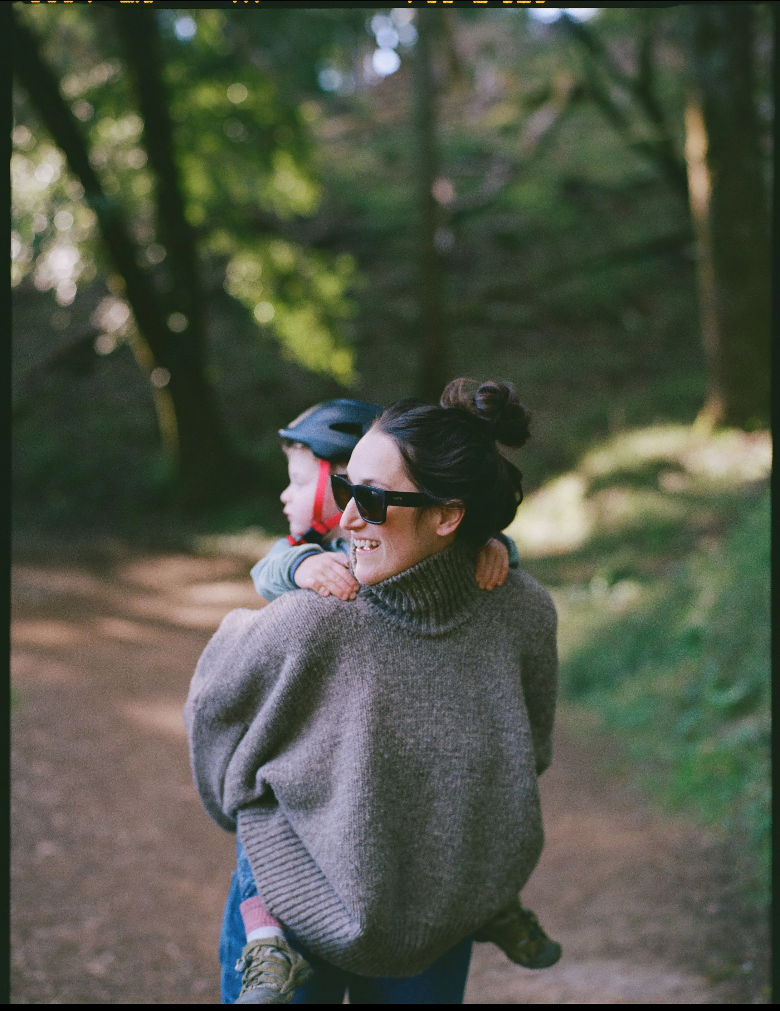 a color medium format film image of a mother giving her 3 year old son a piggy back ride through a wooded area, they are looking off camera. The photo was takin in Marin County 