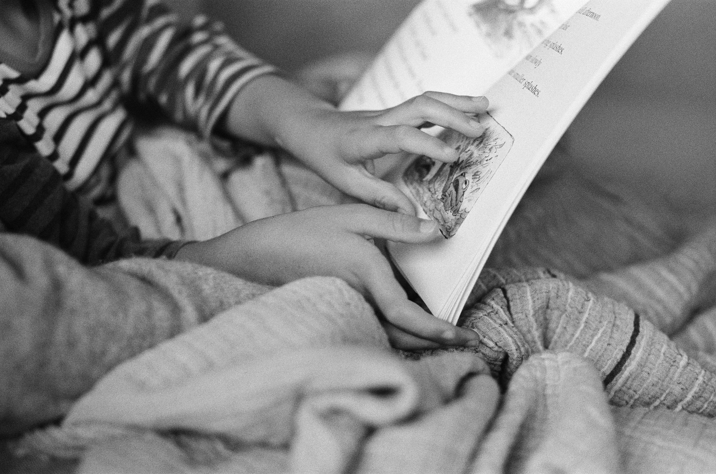 a 35mm black and white photo of a young boy's fingers touching the pages of a book while he reads to his mother who you can't see in the frame. The photo was taken in their home in Sausalito, CA 