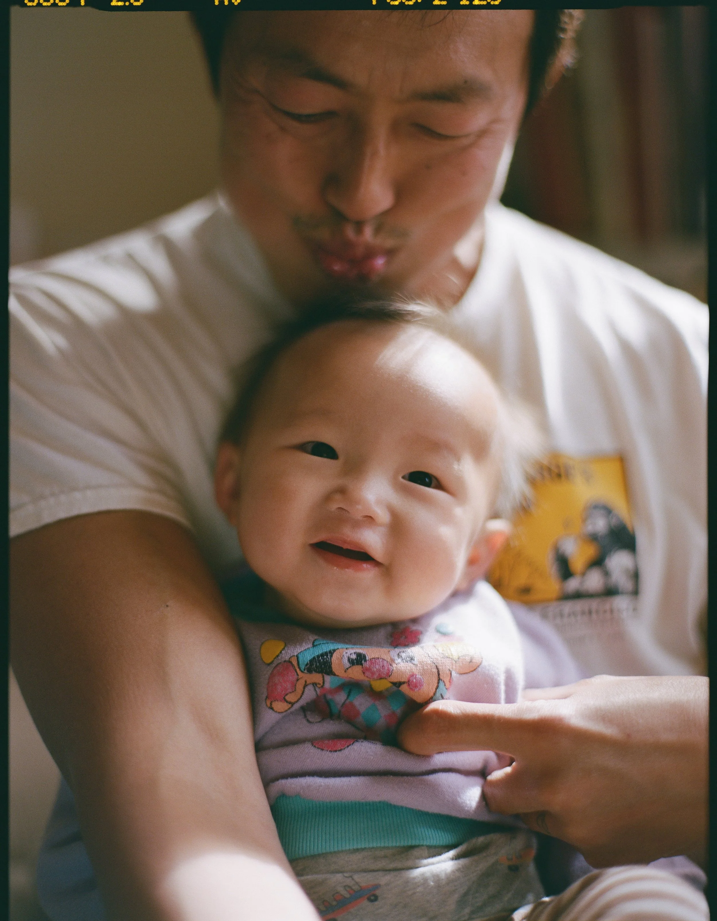 a color medium format film photo of a father holding his infant son in his chest. His son is facing the camera smiling. The photo was taken in their home in San Francisco, California. 