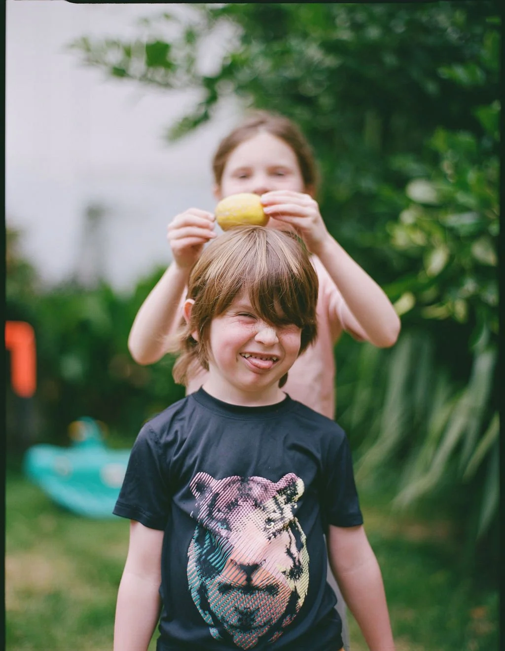 Two children outdoors, one standing in front with a lion graphic T-shirt, sticking out tongue, squinting, and smiling. The other behind, holding a yellow object above the child's head. Green trees in background.