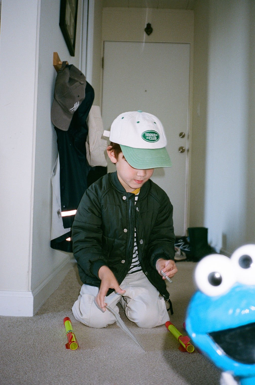 A young boy wearing a white and green baseball cap with a tennis club logo, a black jacket, and light pants kneeling on the carpeted floor indoors. He is holding objects and surrounded by toys. In the foreground, a Sesame Street character, Cookie Mon