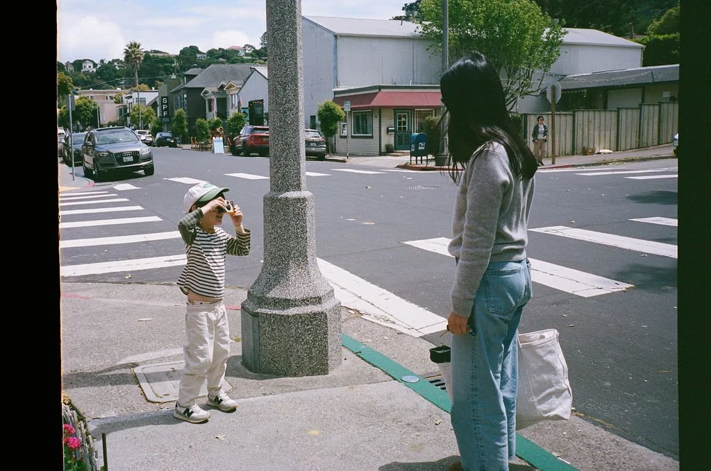 A young child taking a photograph of an adult woman standing at a crosswalk on a city sidewalk.