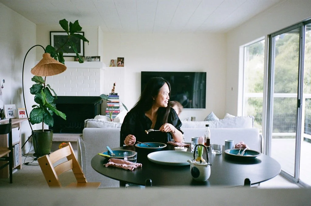 Woman sitting at a dining table in a bright living room, with sliding glass doors and outdoor scenery visible through the windows.