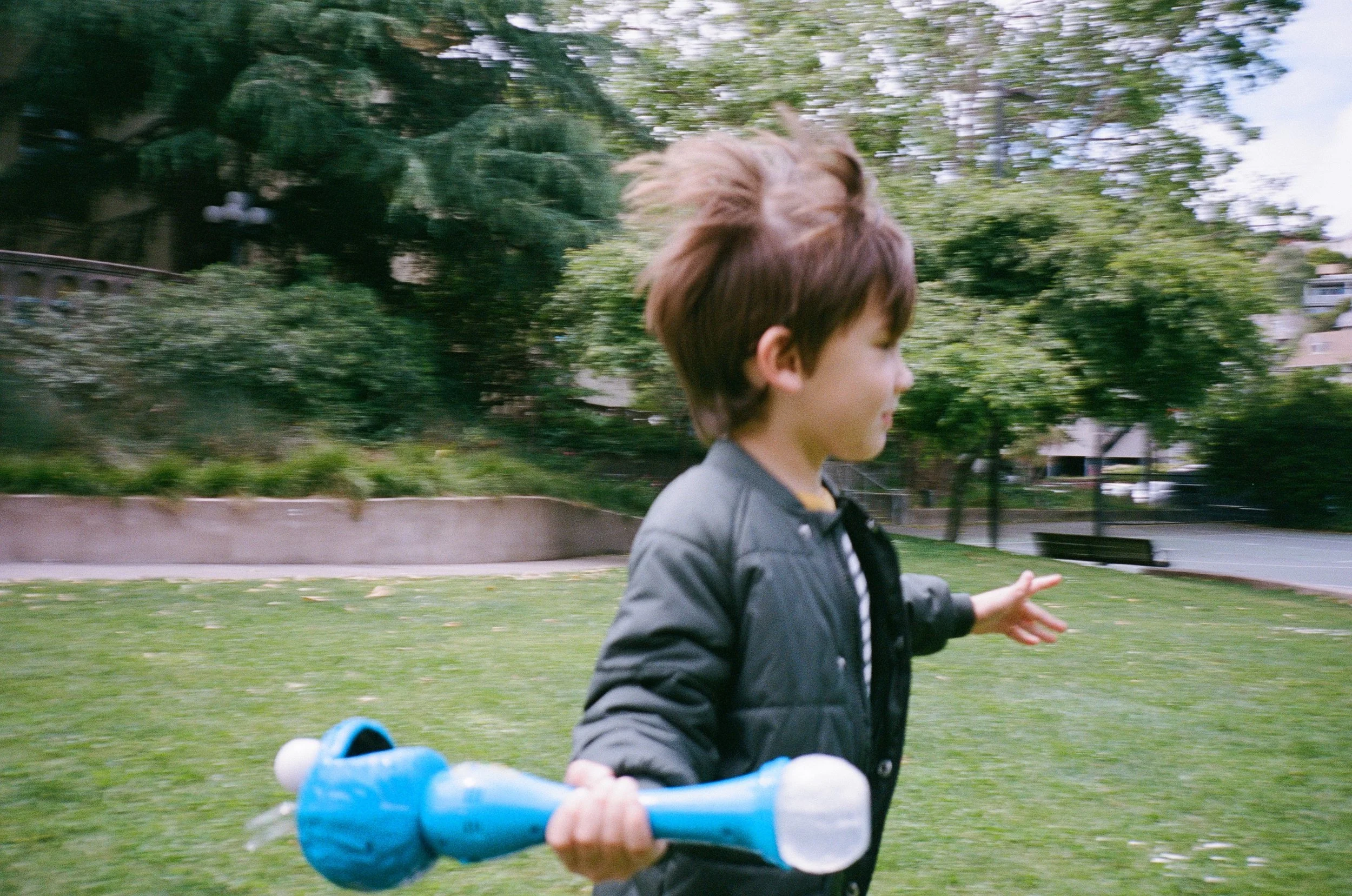a color 35mm film photo of a little boy with brown hair running in a park holding a bubble wand that looks like Cookie Monster from Sesame Street, the photo is blurry to convey motion. The photo was taken at a park in Sausalito, California 