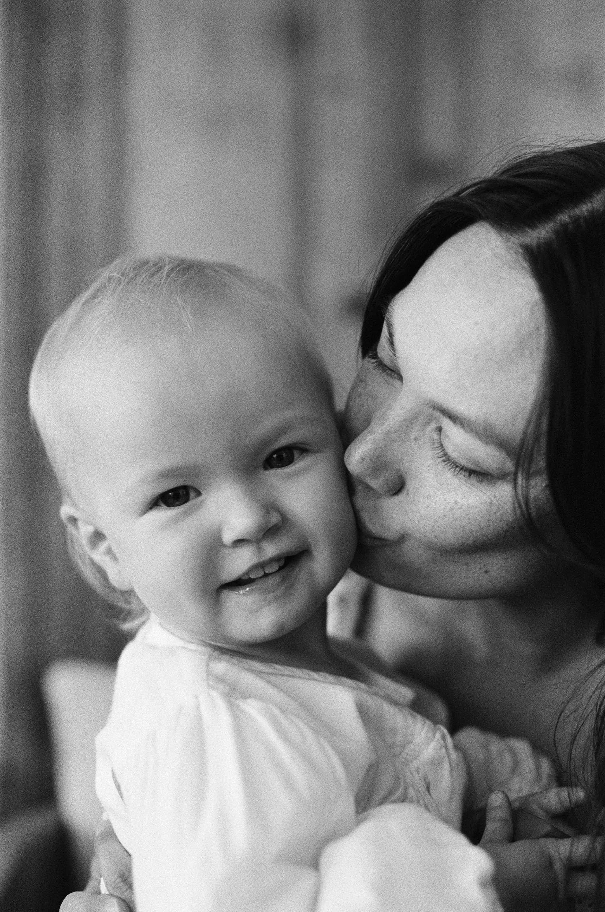 a black and white 35mm film photo of a mother kissing the cheek of her infant daughter who is looking at the camera in their home in Sausalito, California 