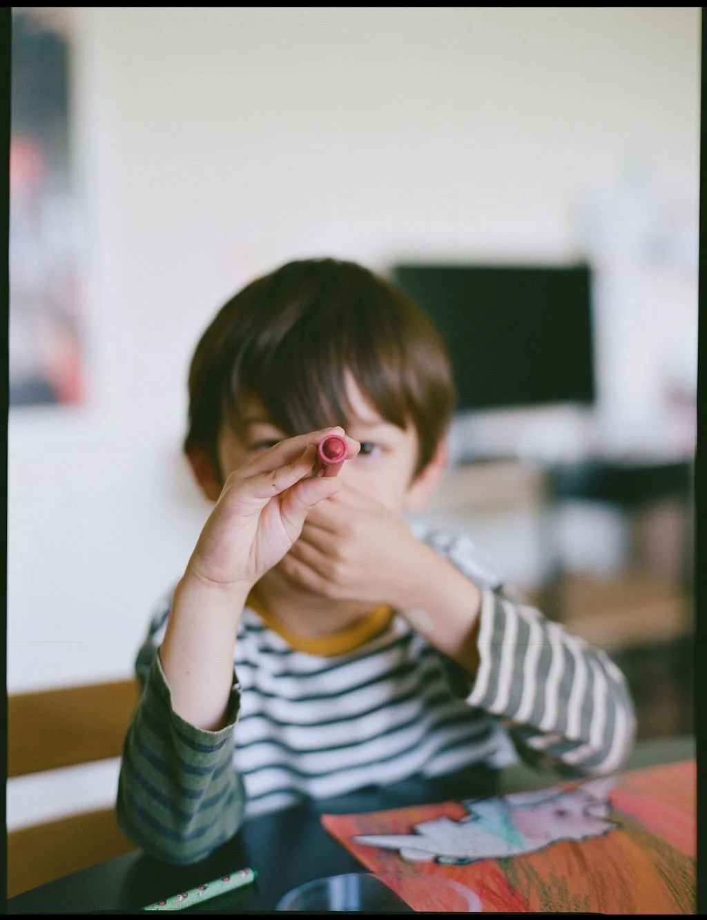 A young boy with brown hair wearing a striped shirt, holding a pink crayon to his eye while sitting at a table with colorful drawings and papers.