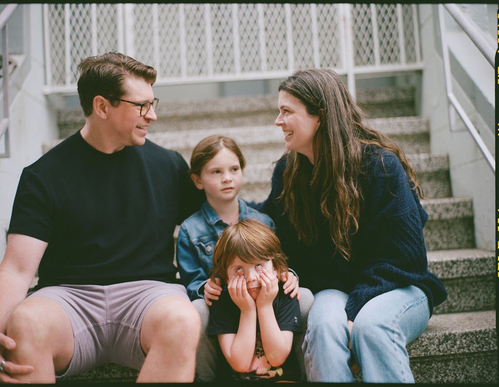 A family of four sitting on outdoor stairs, smiling and talking. The father is wearing glasses and a black t-shirt, the mother has long brown hair and a navy sweater, the daughter has brown hair and a denim jacket, and the son has red hair and is res