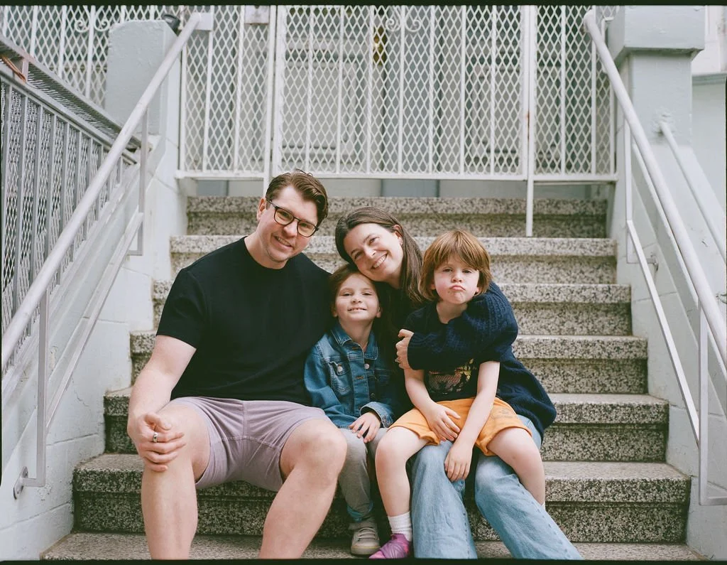Family of four sitting on outdoor stairs, smiling at the camera. The father has glasses and wears a black t-shirt and shorts. The mother has dark hair and wears a black top. The young girl has a denim jacket, and the young boy has a navy sweater and 