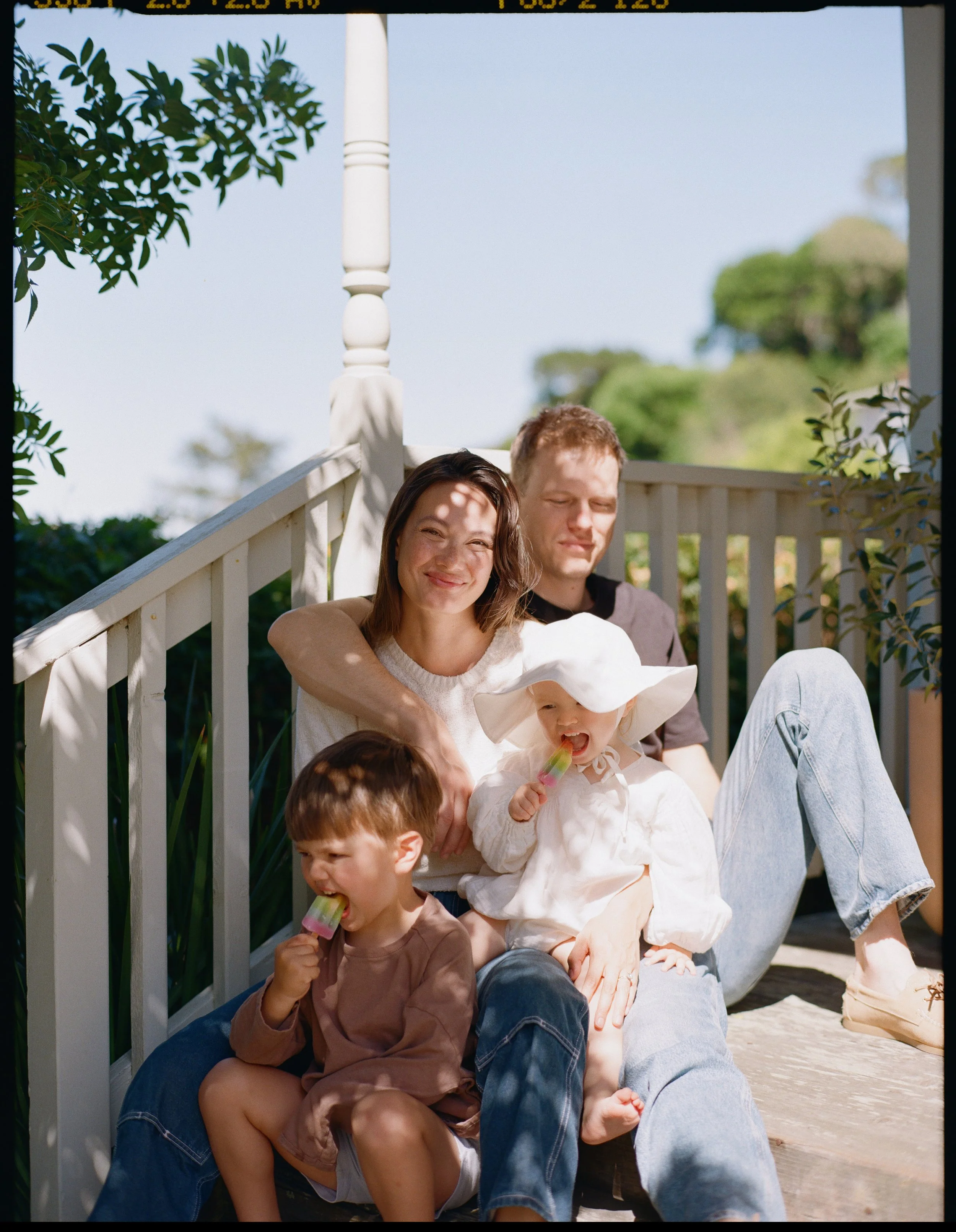 color medium format film image of a family of 4 on the front steps of their house in Sausalito, California. The light is dappled on their faces and the little boy and little girl are eating popsicles. 