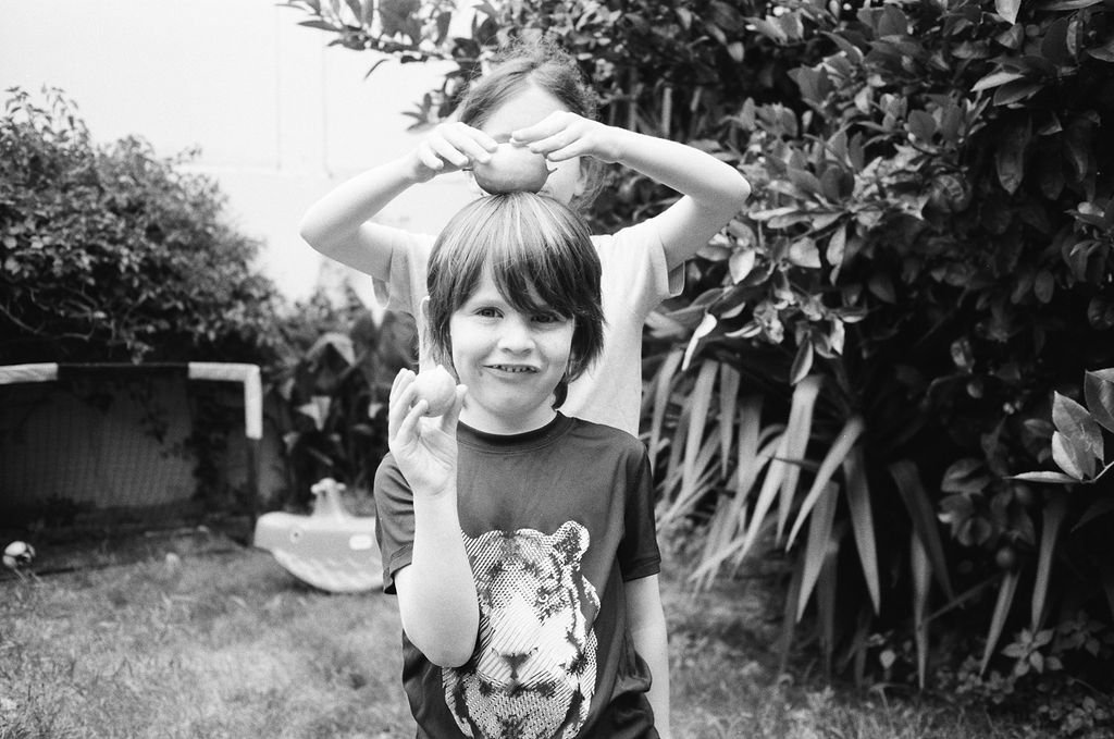 Two children in a garden, one holding an apple on top of his head and smiling, the other holding an apple in front, with greenery in the background.