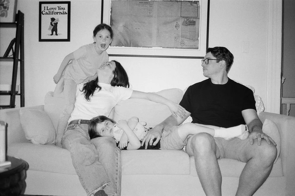 A woman is sitting on a sofa with two young children, one lying on her lap and the other standing behind her, both reaching out toward a man sitting on the sofa to her right. The woman appears to be playfully reaching for the child standing. The man 