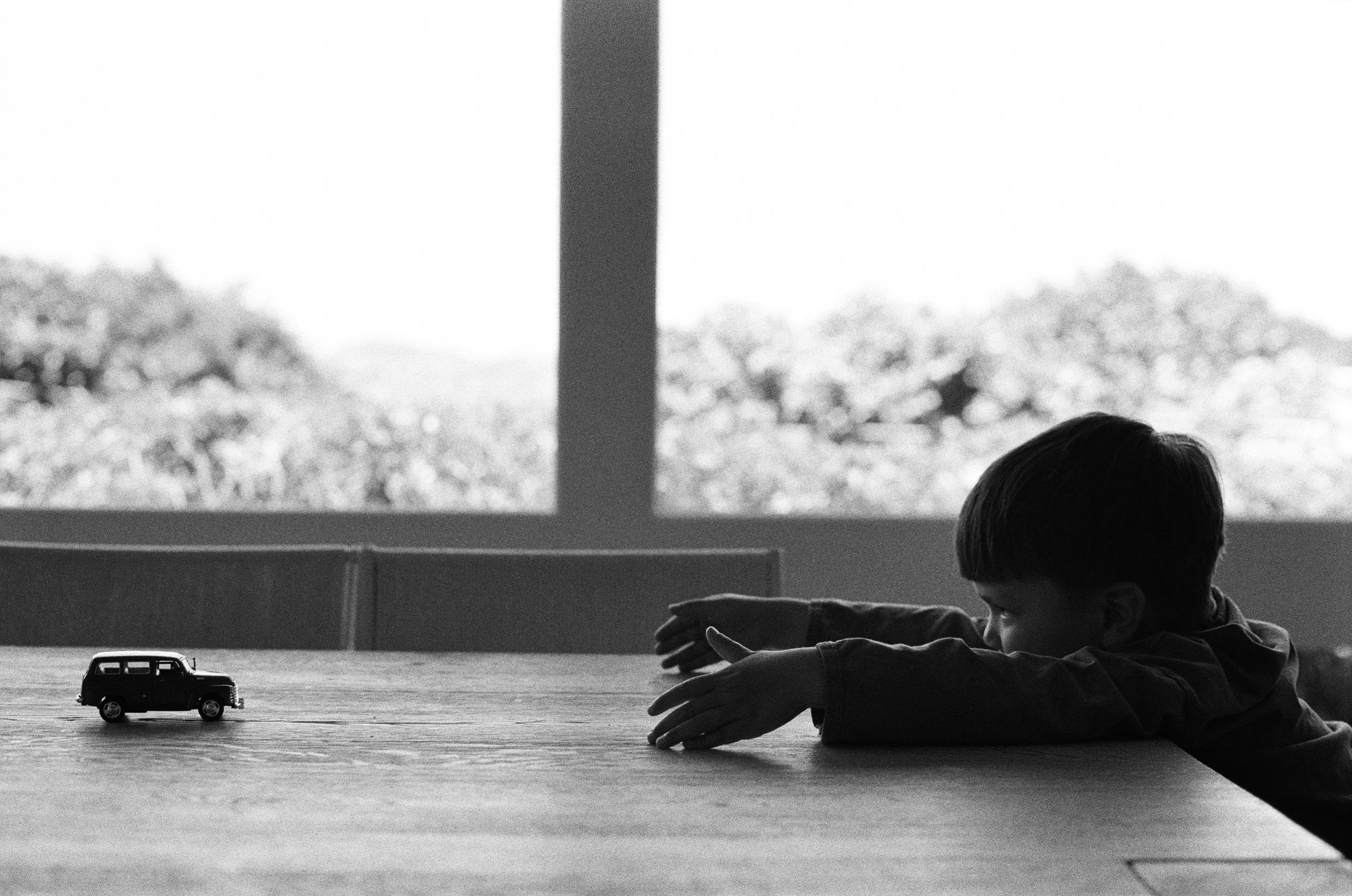 a black and white 35mm photo of a little boy sitting at the dinner table with his arms stretched out reaching for a little toy car that is just out of his reach. The photo was taken at their home in Sausalito, California 