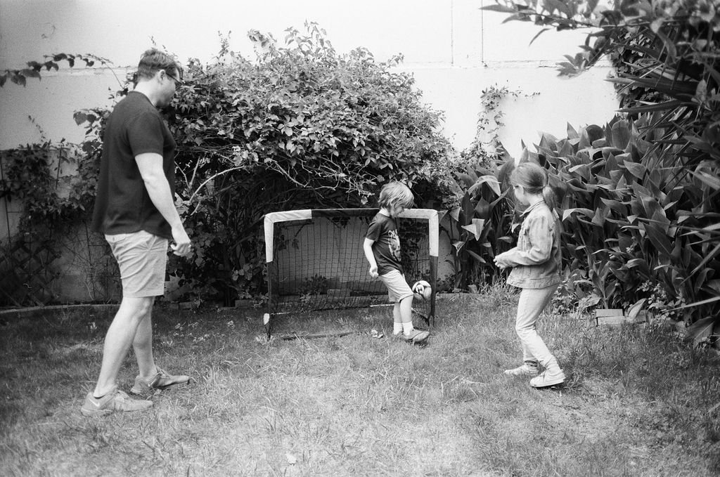 A man and two children playing soccer in a backyard with a small goal, surrounded by bushes and trees.