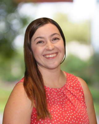 A young woman with long brown hair smiling, wearing a red sleeveless dress with white polka dots, outdoors with blurred green trees in the background.