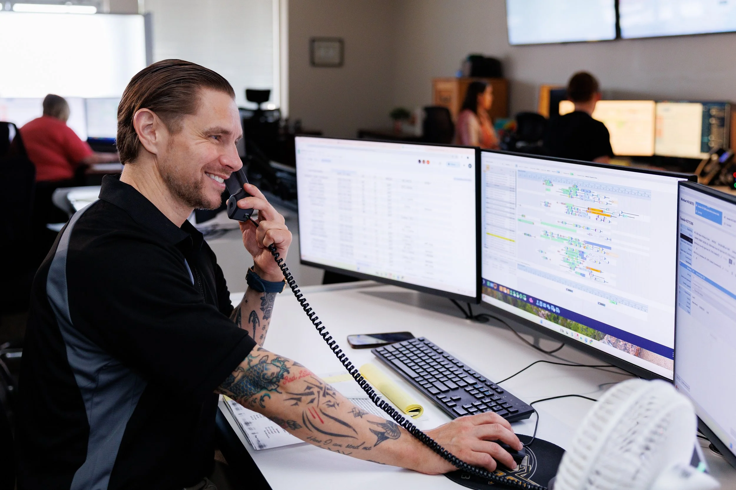 A man with tattoos on his arms is smiling while talking on the phone at a desk with dual monitors displaying colorful data charts and spreadsheets in a busy office environment.