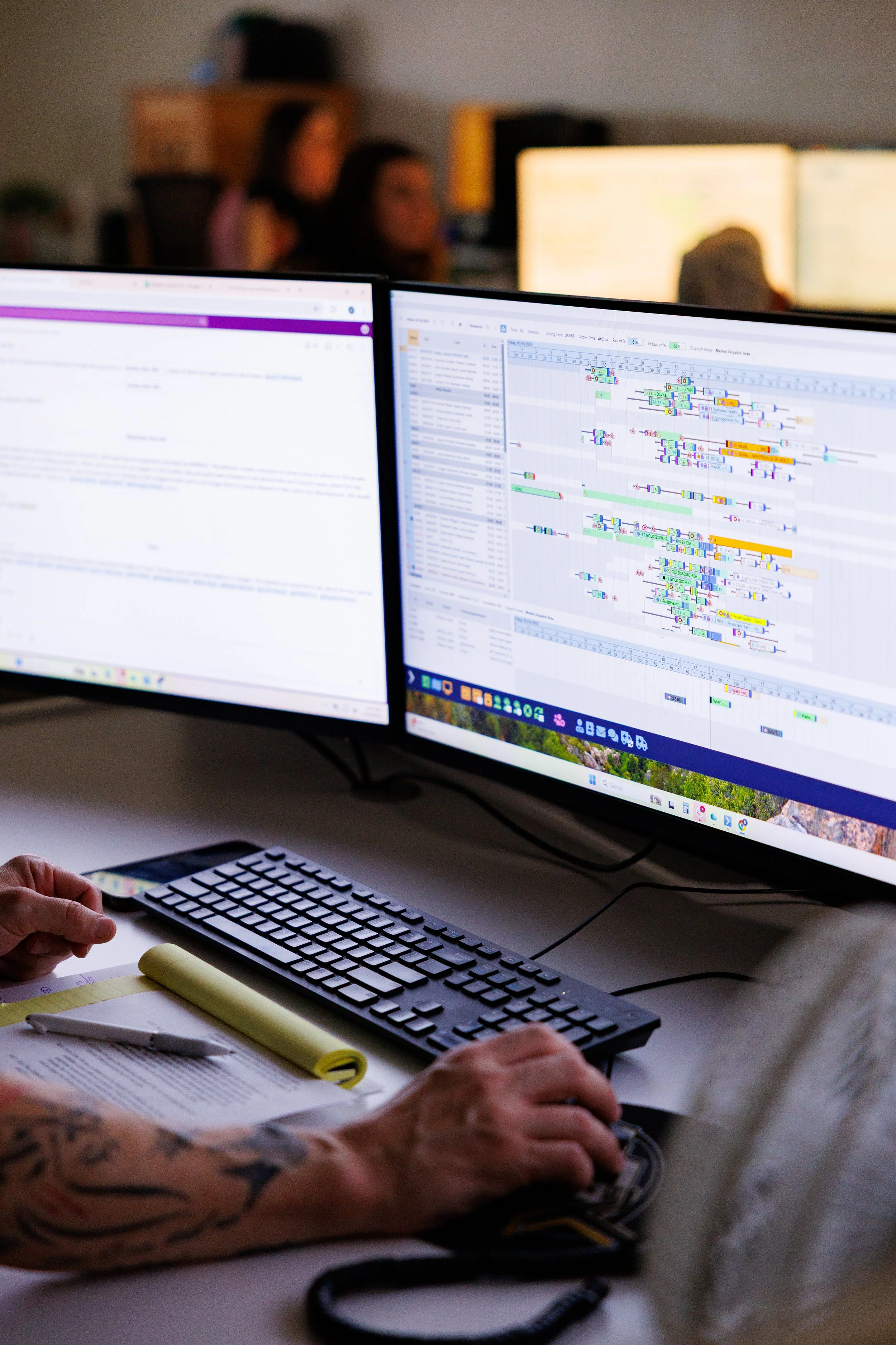 Two computer monitors displaying software with charts and data. A person with tattooed arms is working with a keyboard and mouse in front of the monitors. There are notebooks, papers, and a pen on the desk.