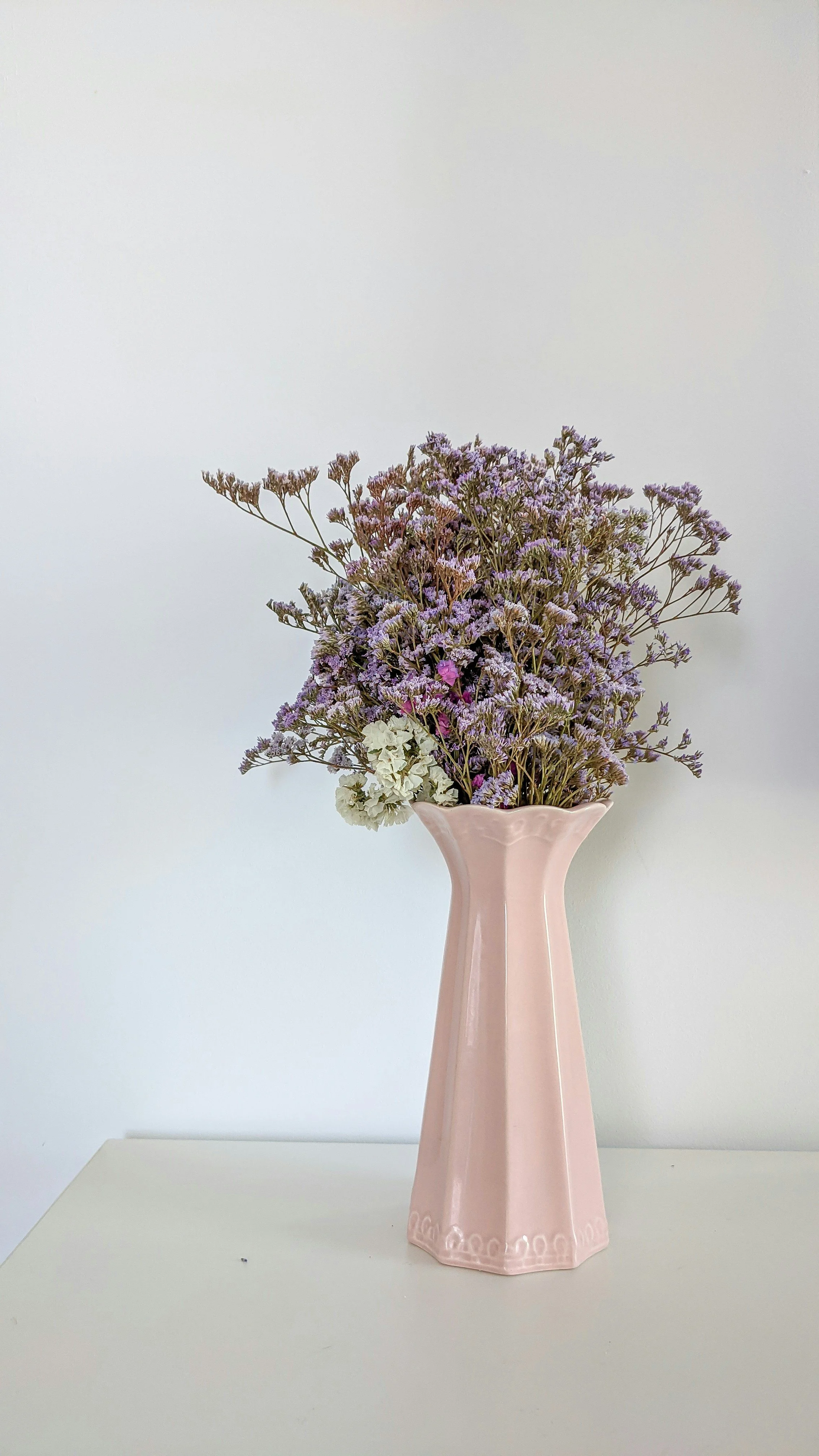 Pink ceramic vase with white and purple flowers on a white surface against a white background.