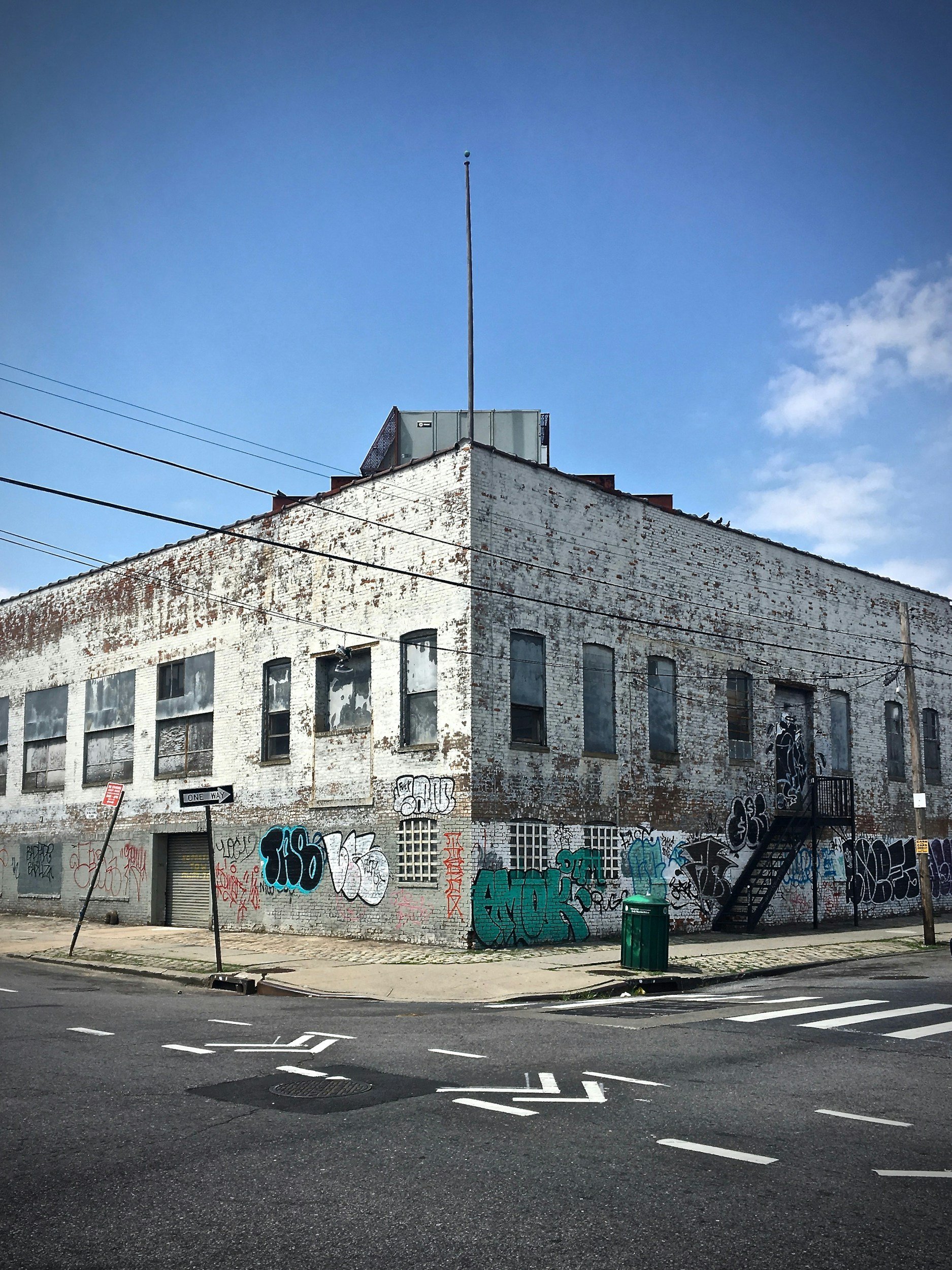 A corner of an old brick building with graffiti on its walls, metal fire escape ladder, and a green trash bin on the sidewalk under a partly cloudy sky.
