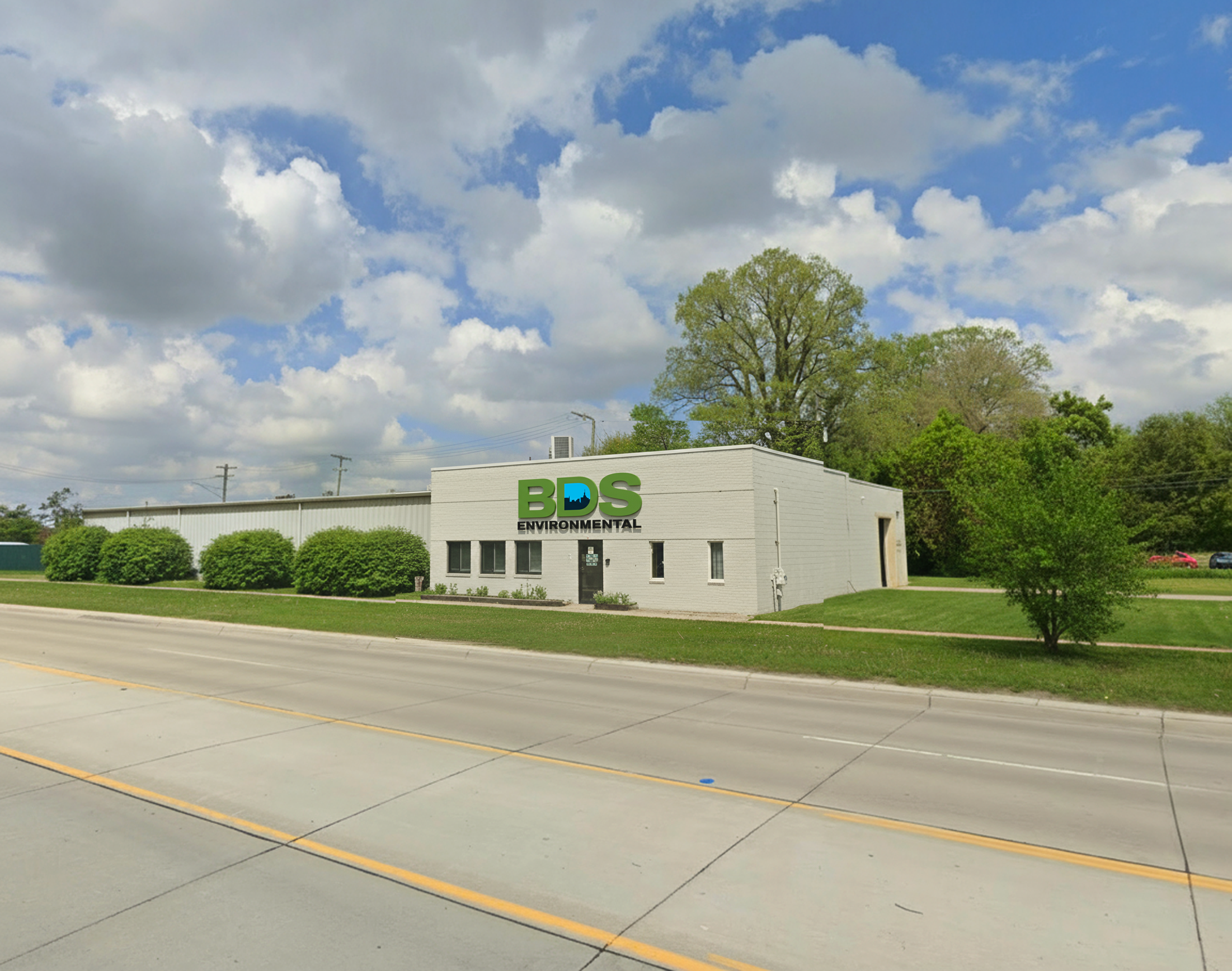 A white building with a sign that reads 'BDS Environmental' in green and black, located beside a road with green bushes and trees in the background, under a partly cloudy sky.
