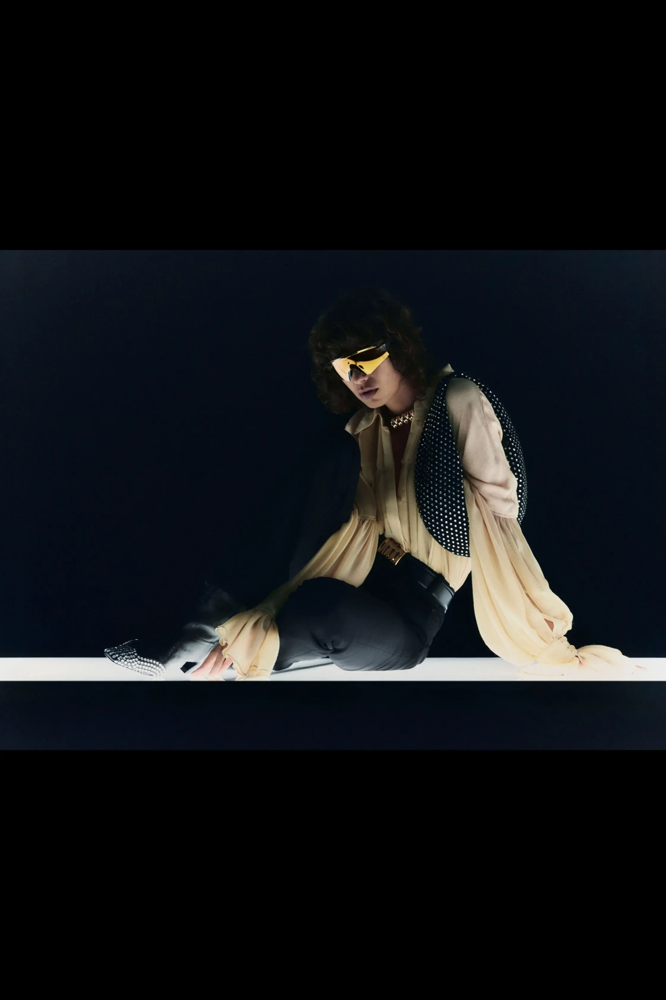 A woman with curly hair wearing yellow futuristic glasses, a beige flowing blouse, black pants, and a black and white polka dot vest, is sitting on a white surface against a black background.