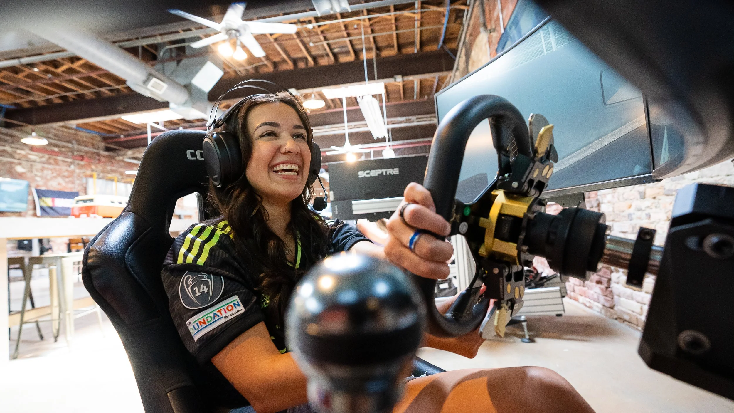 Young woman with long dark hair smiling while sitting in a racing simulator, wearing large headphones and holding a racing wheel in a sim racing center with exposed brick walls and wooden ceilings.