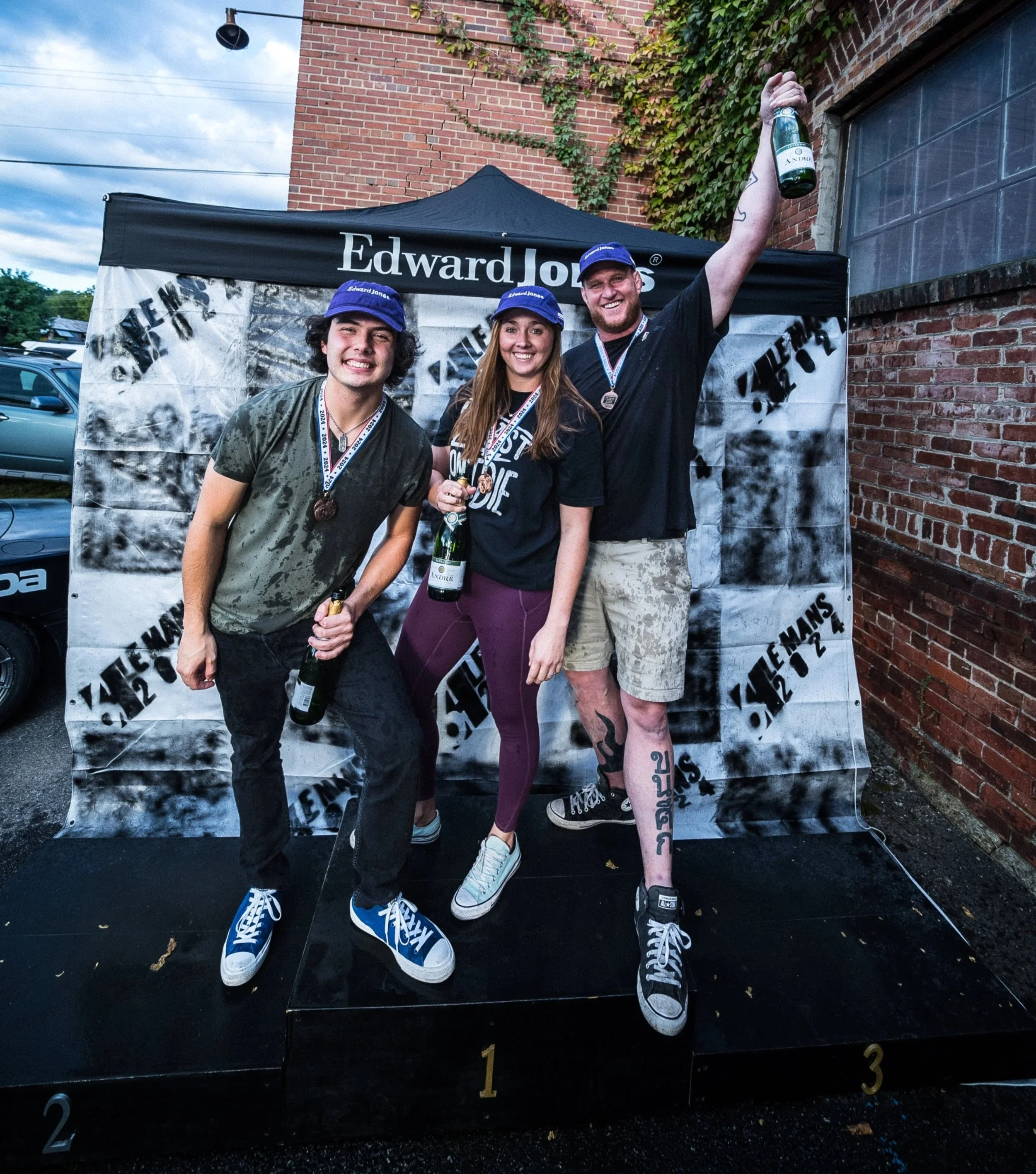 Three young people celebrating on a winners' podium with medals and bottles of champagne, under a black and white tent after a 6 hour endurance race, outside near a brick wall.