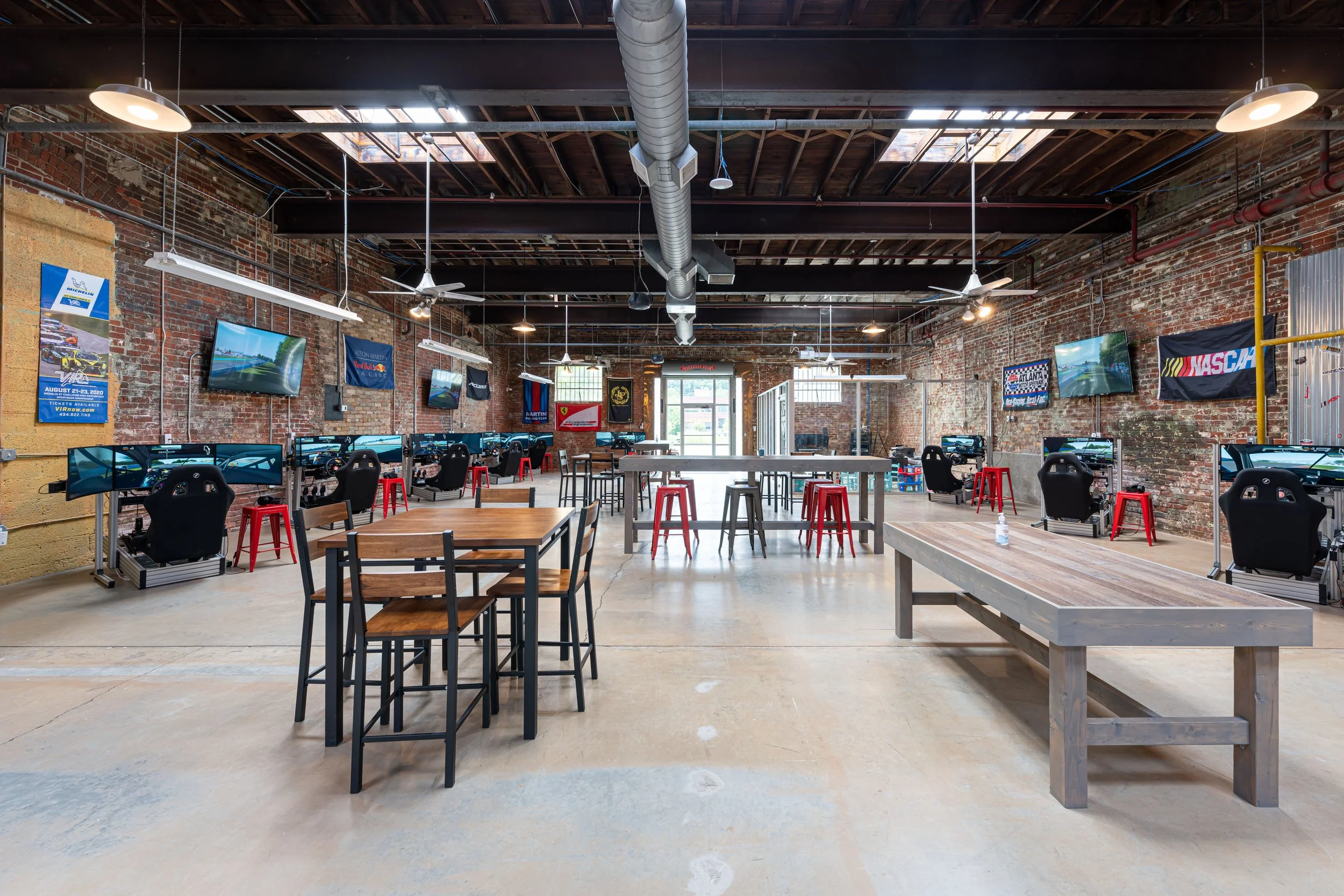 Sim racing center interior with multiple Tvs along the walls, exposed brick walls, and flags, including NASCAR, lining the walls. There are several chairs, tables, and stools, with a mix of concrete and wooden surfaces.