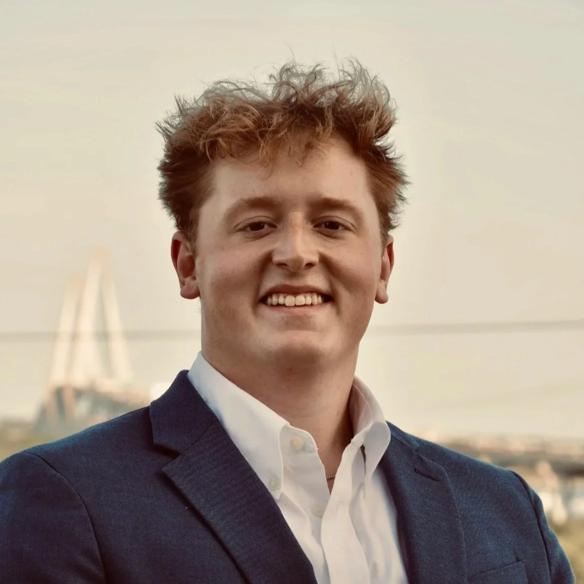 Portrait of a young man with red hair, wearing a white shirt and dark blazer, smiling outdoors with a blurred body of water and bridge in the background.